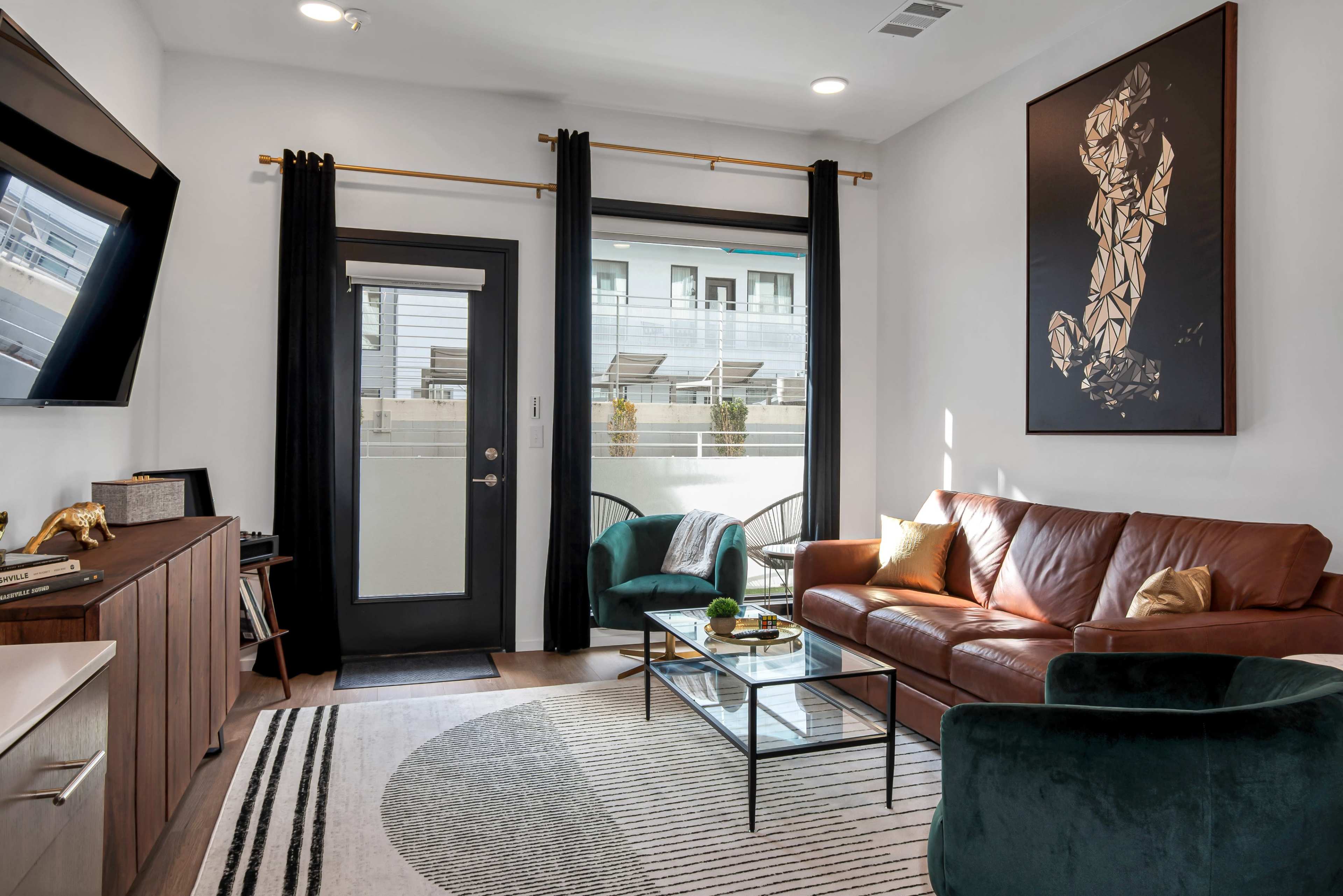 A modern living room with a brown leather sofa, a glass coffee table, and a door leading to a balcony, complemented by black curtains and a large artwork on the wall.