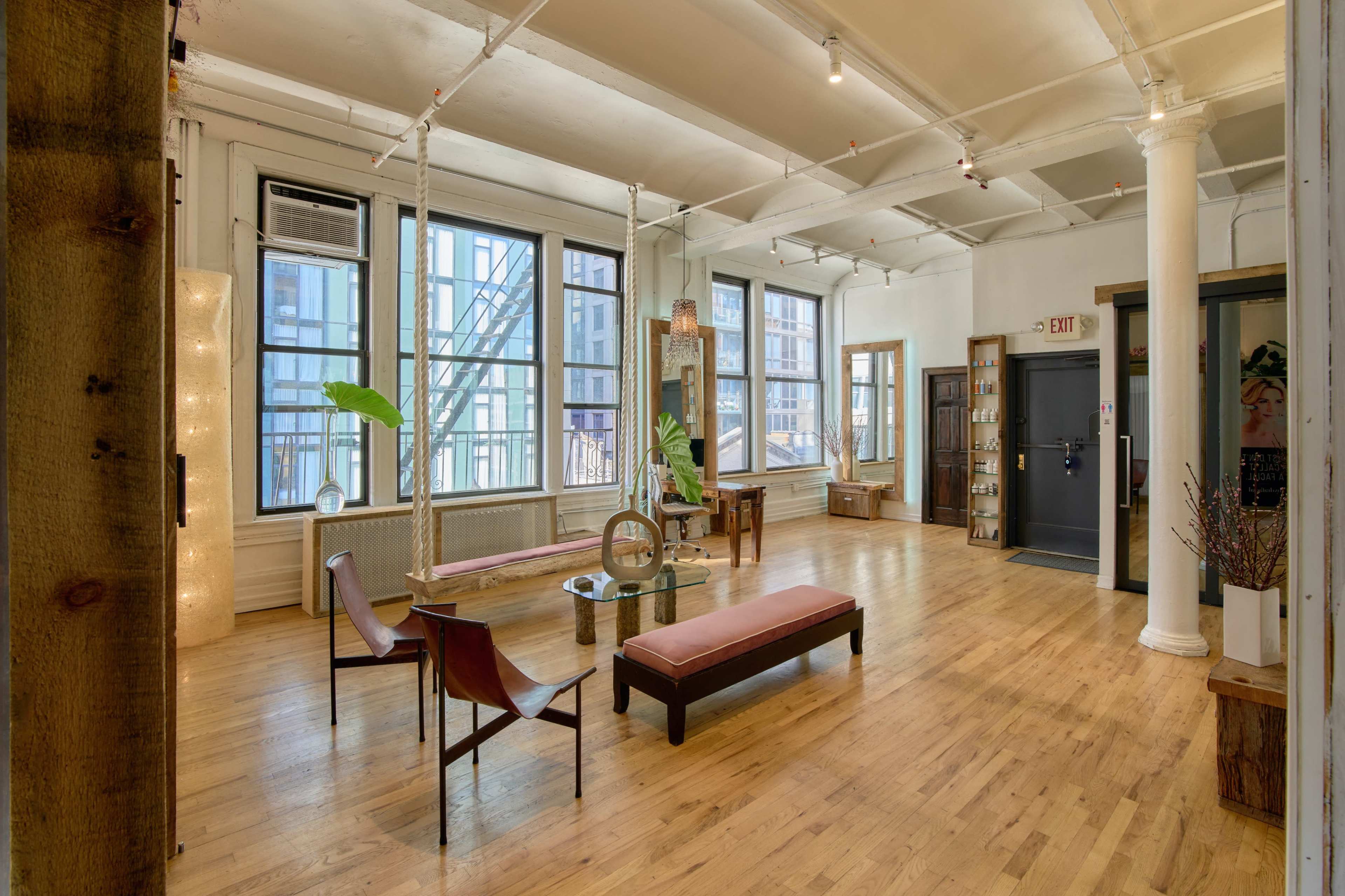 The image shows a spacious, well-lit room with wooden floors, featuring two chairs, a long bench, a small table, and large windows along one wall.