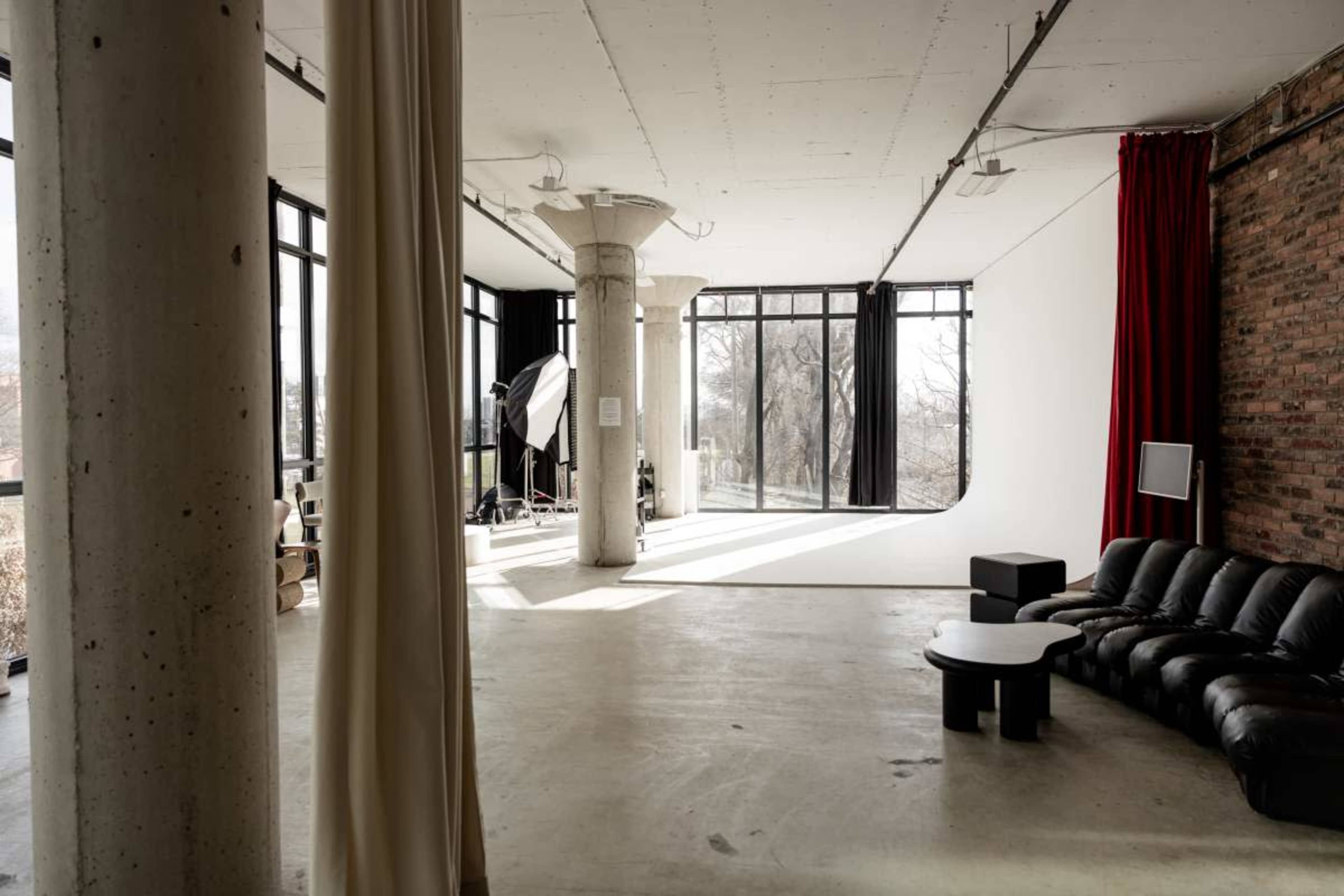 The image shows a spacious, modern studio with large windows, exposed brick walls, a black leather sofa, and various lighting equipment in the background.