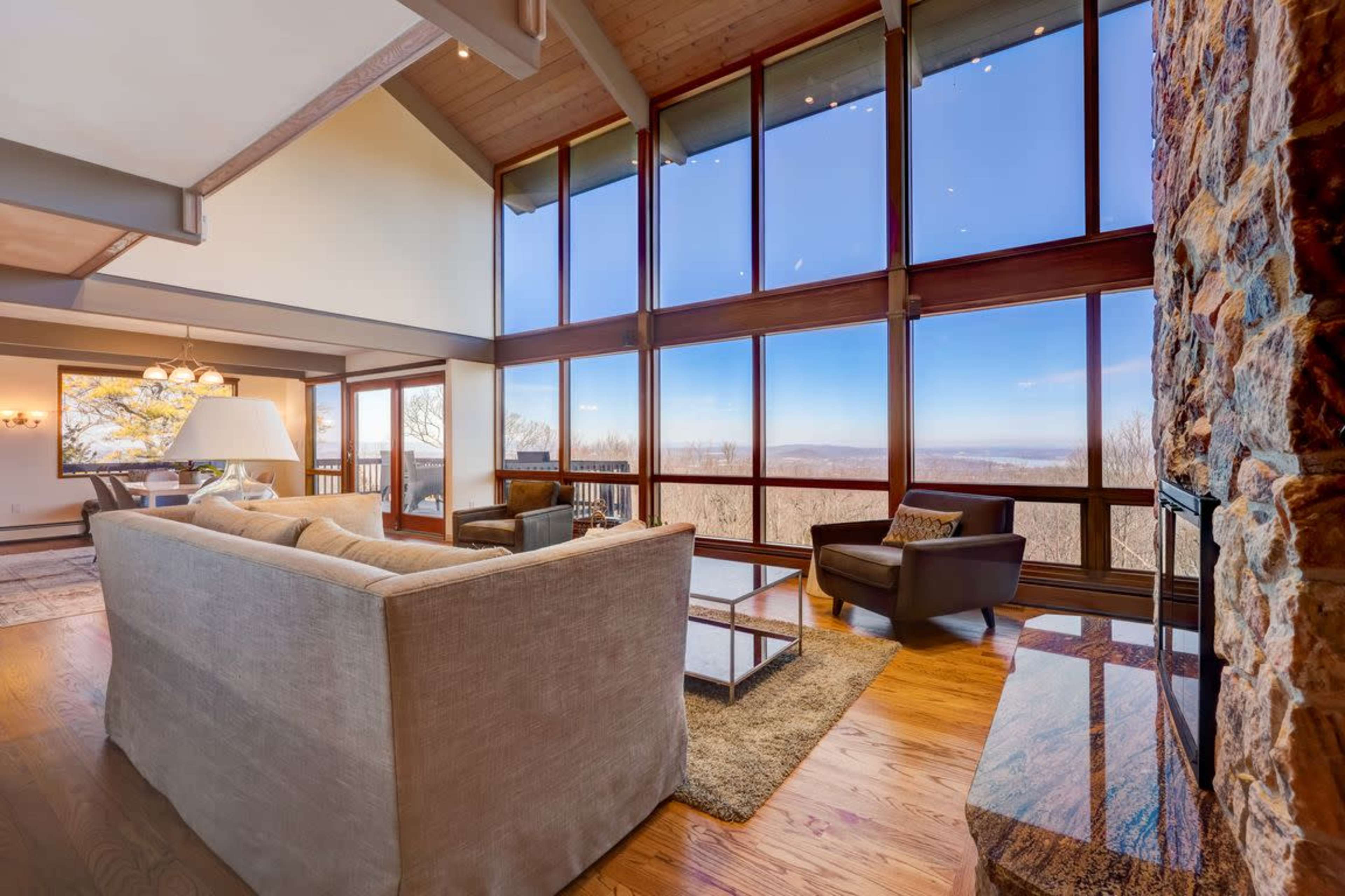 A spacious living room features large windows overlooking a scenic view, with modern furniture arranged around a coffee table and a stone fireplace.
