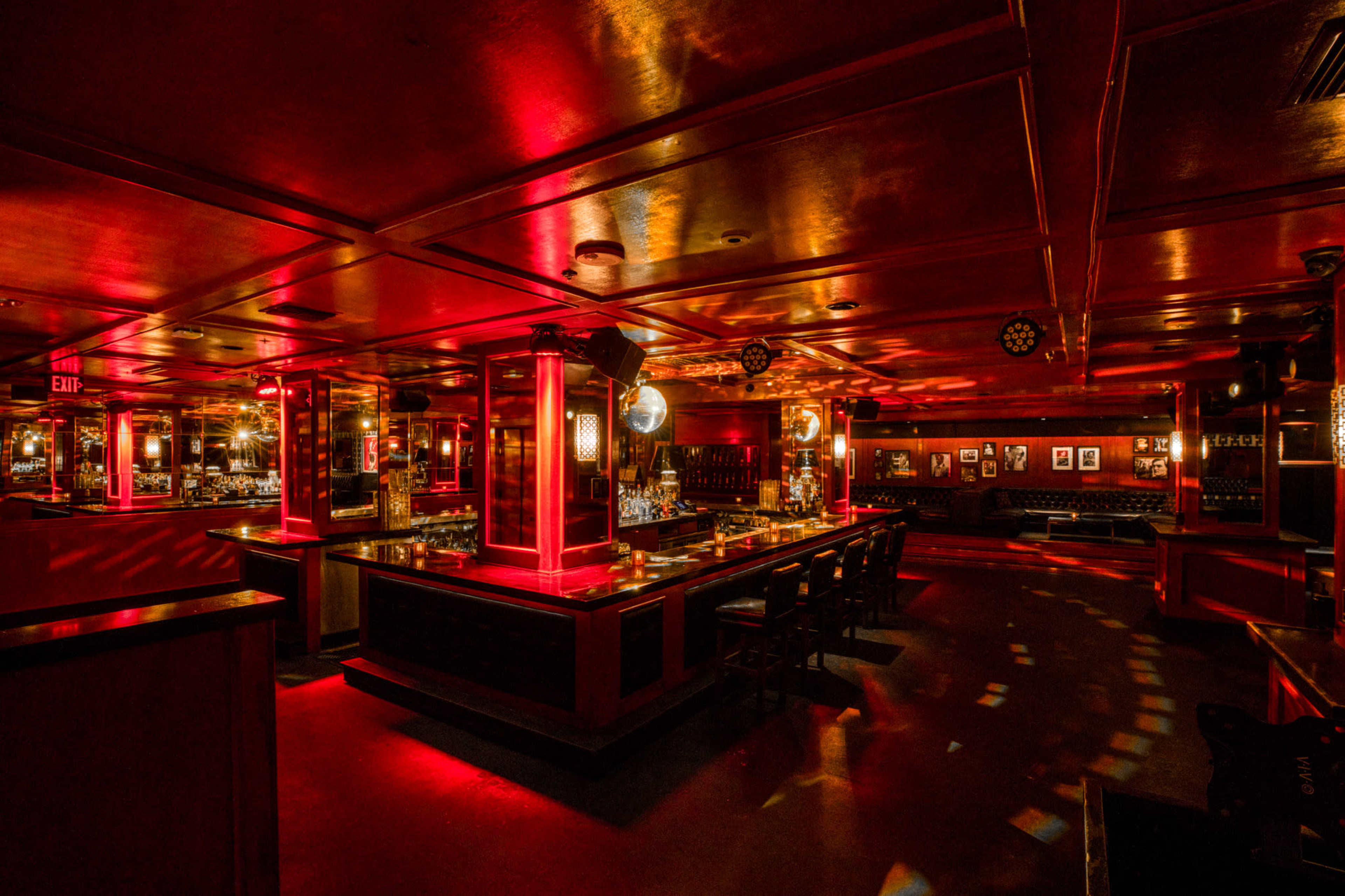A dimly lit bar interior with red lighting, a long bar counter, and circular tables surrounded by chairs.
