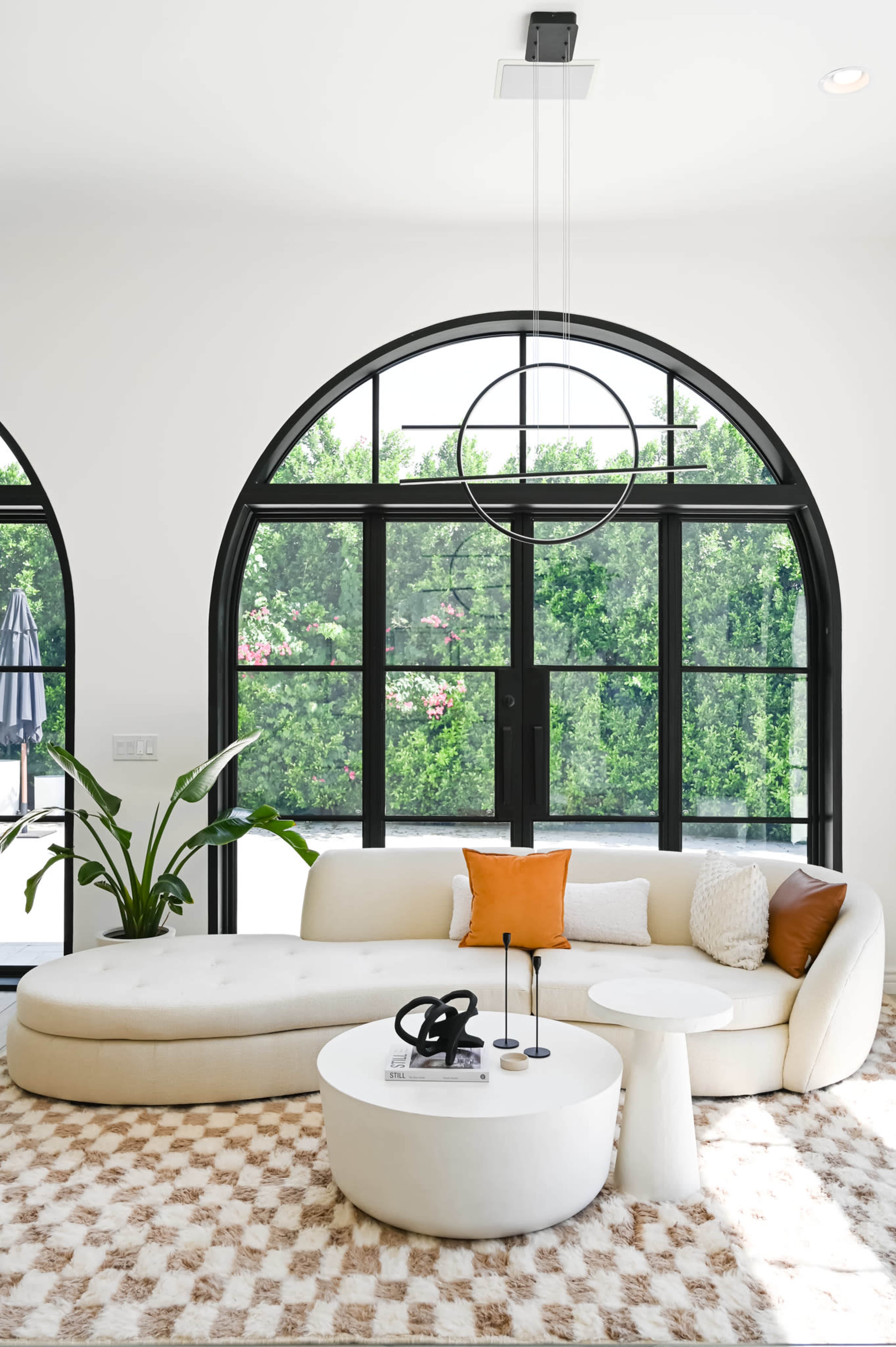 A modern living room features a curved white sofa, a round coffee table, and large arched windows with lush greenery outside.