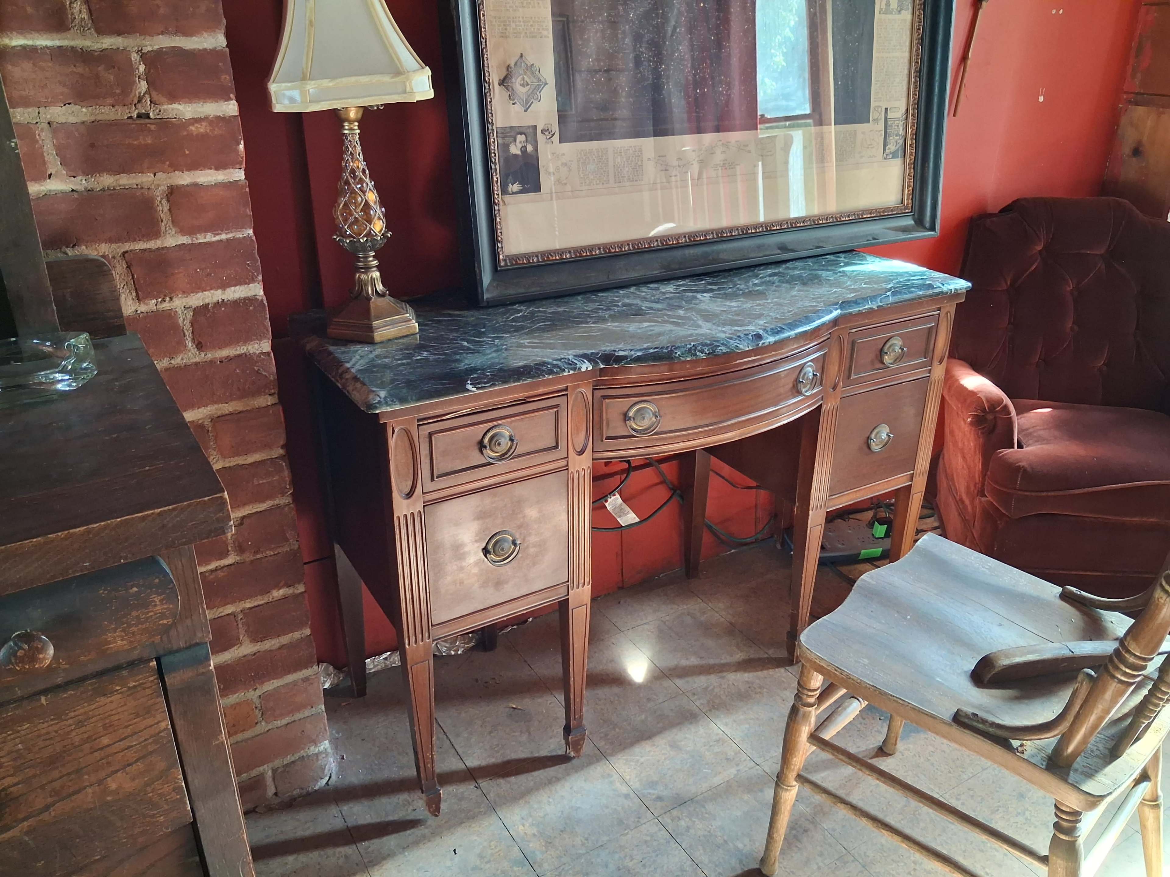 The image shows a wooden sideboard with a marble top, flanked by a lamp and a framed picture against a red wall, alongside a vintage chair and brick wall.