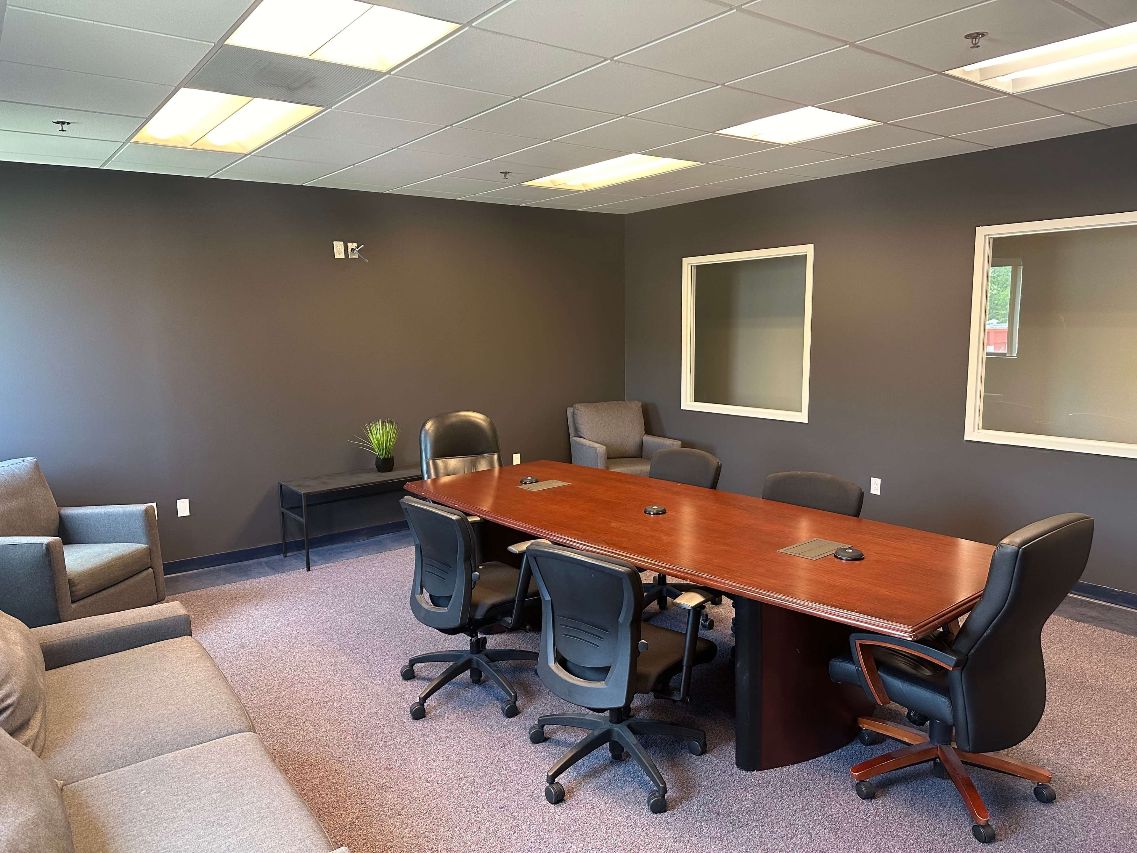 A conference room with a large wooden table surrounded by black chairs and a small seating area with gray sofas.