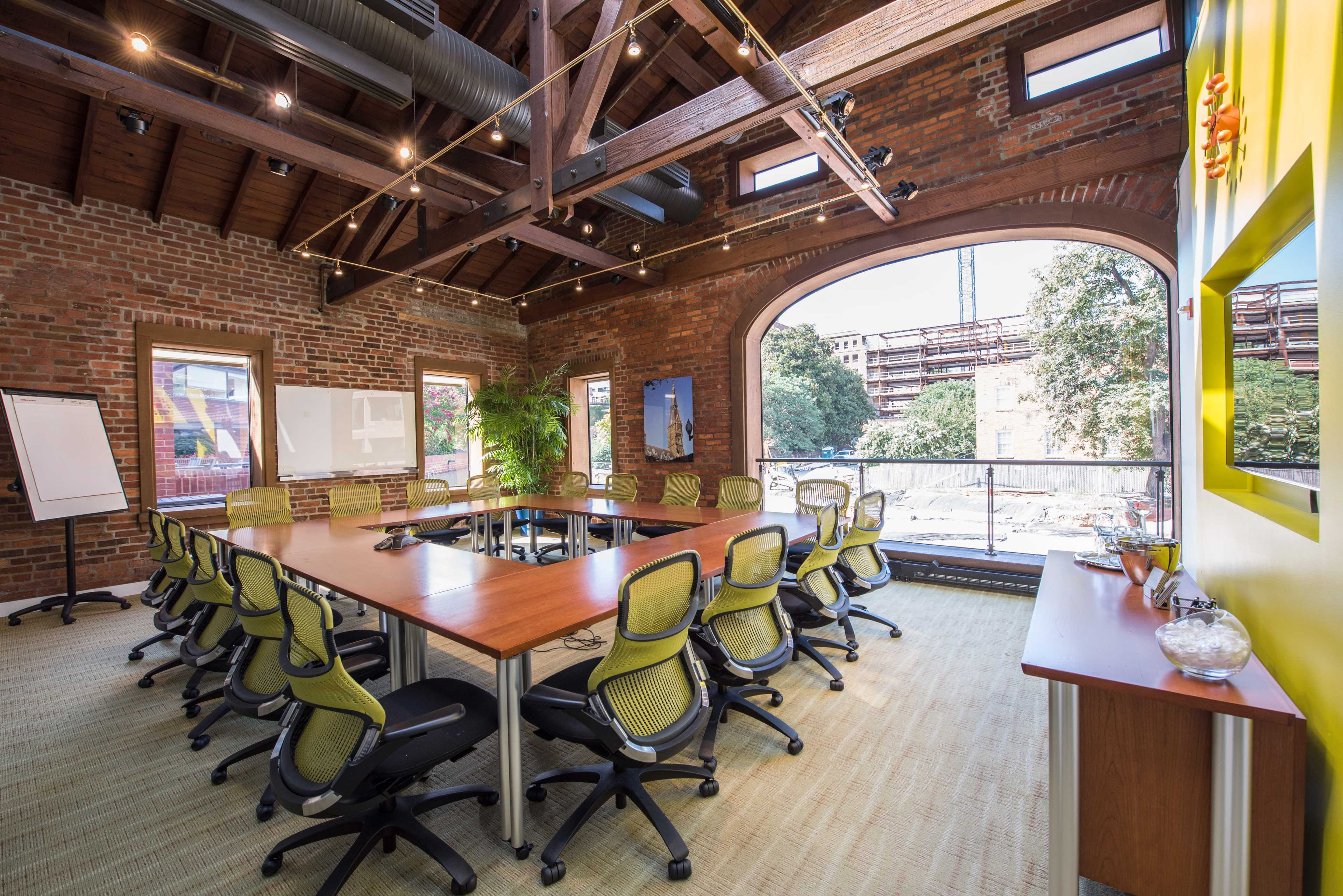 A conference room with a large table surrounded by rolling chairs, set against a backdrop of exposed brick walls and large windows offering a view of greenery outside.