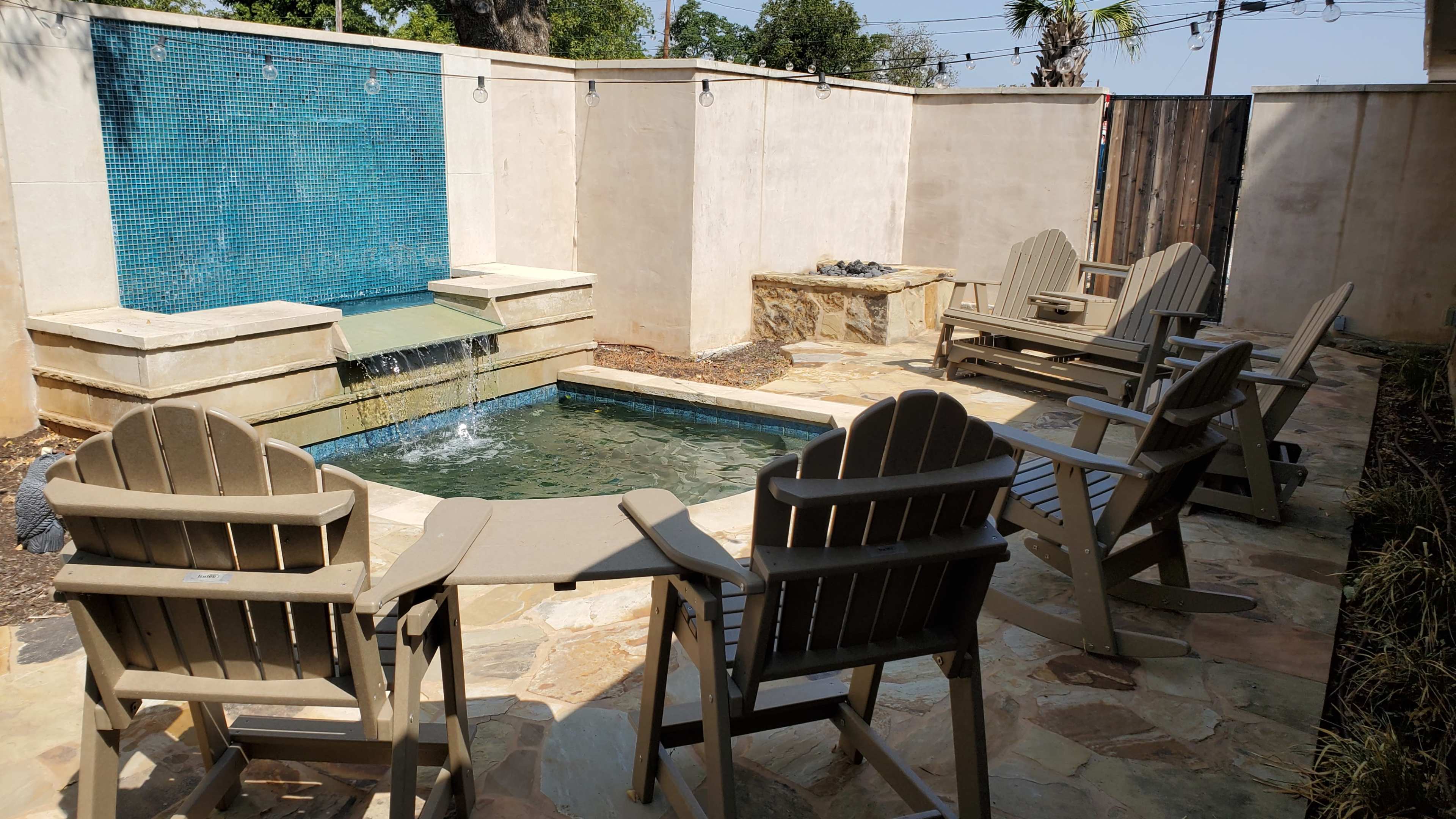 The image depicts a small outdoor area featuring a shallow pool with a water fountain, surrounded by several Adirondack chairs on a stone patio.
