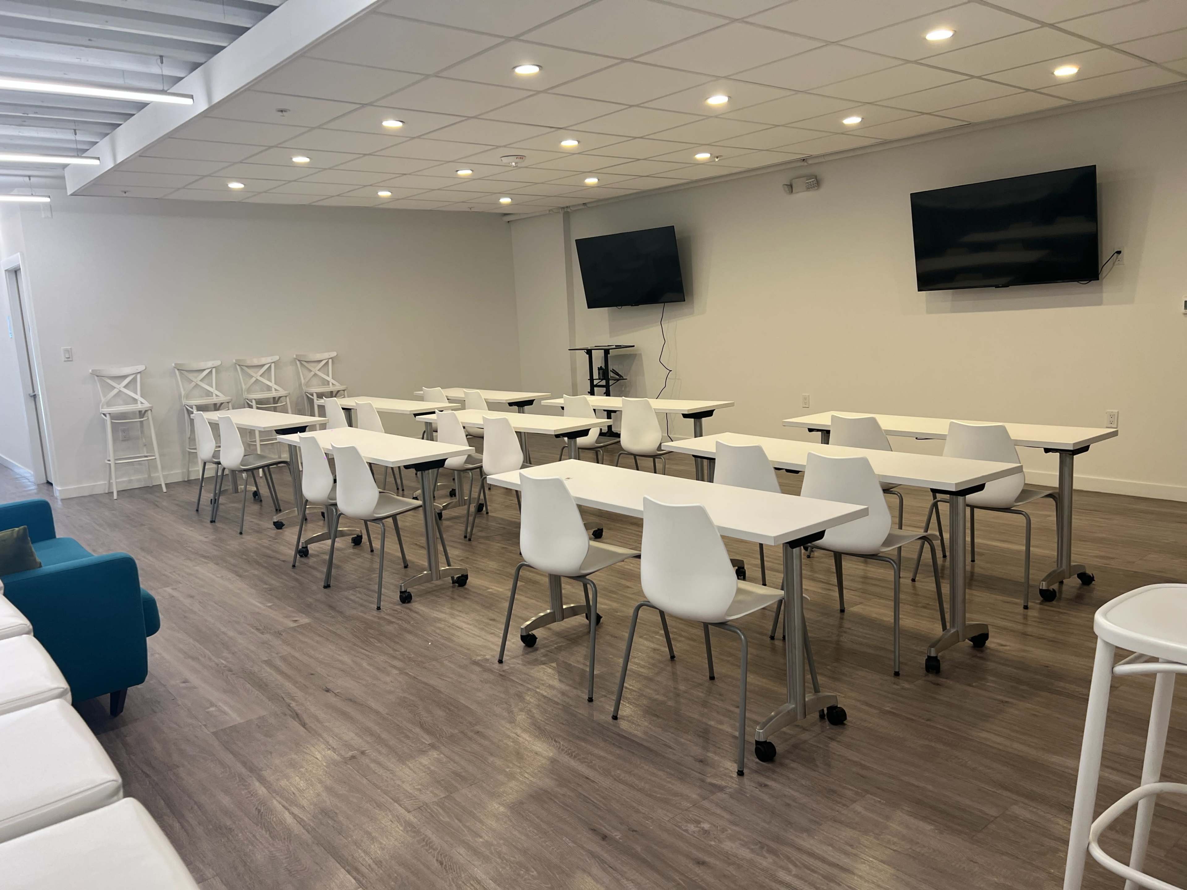 A spacious, well-lit meeting room with multiple rectangular tables and chairs arranged in rows, flanked by two wall-mounted televisions.
