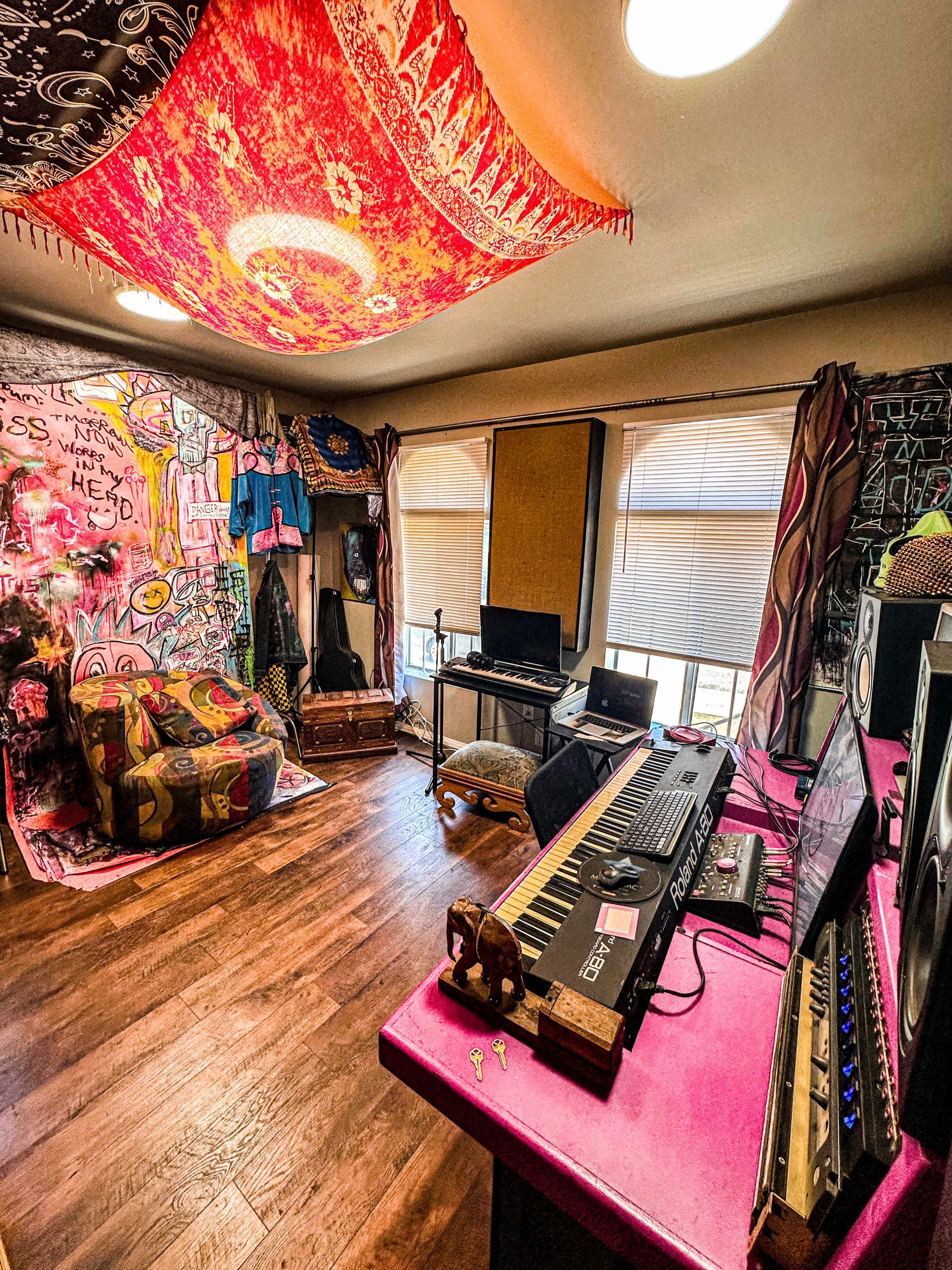 A colorful music studio features a keyboard, computer setup, and a vibrant tapestry hanging from the ceiling, alongside a decorated wall and seating area.