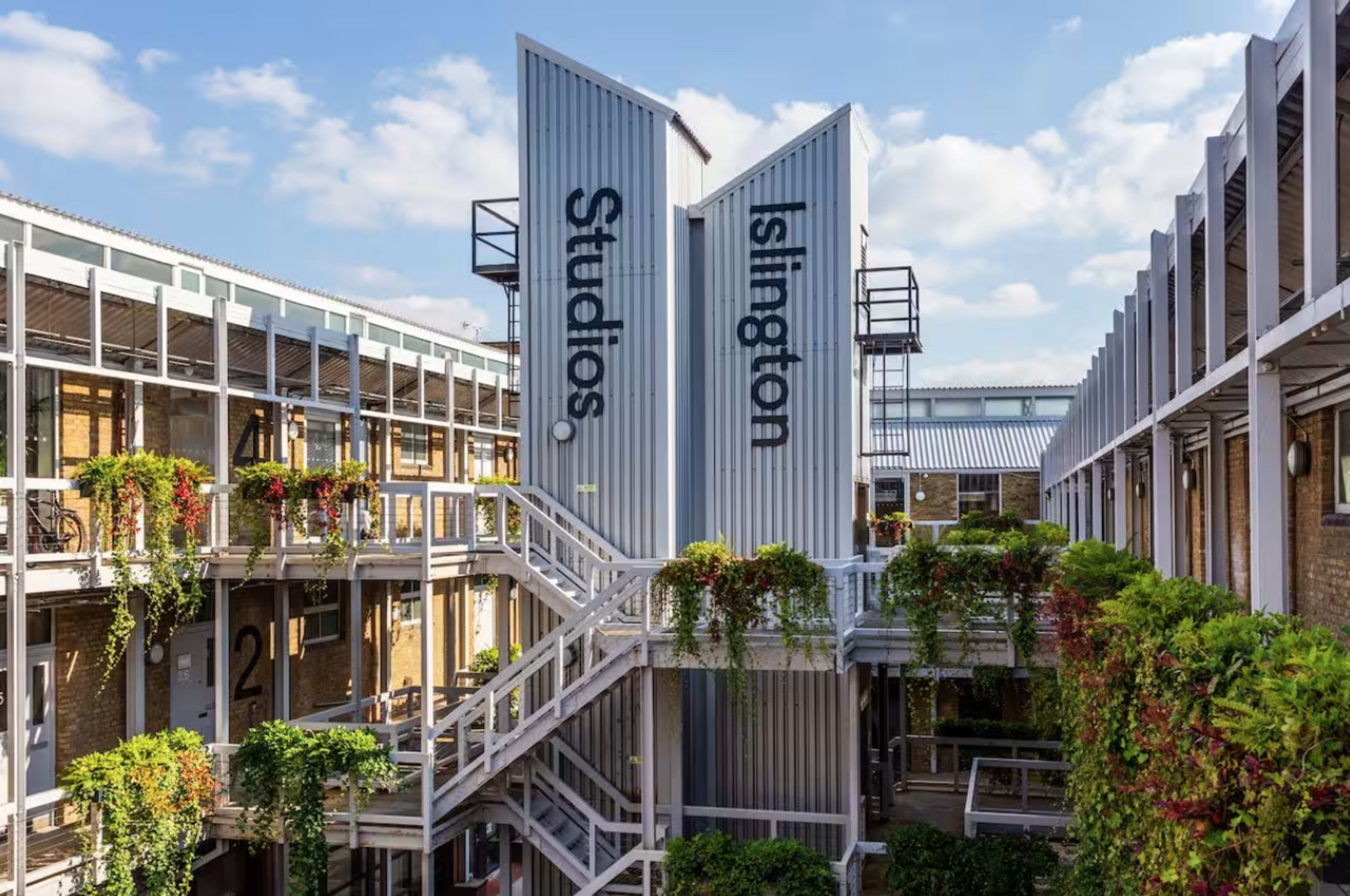 A modern building complex with a courtyard, featuring metal structures labeled "Studios" and "Islington," surrounded by greenery and stairs.