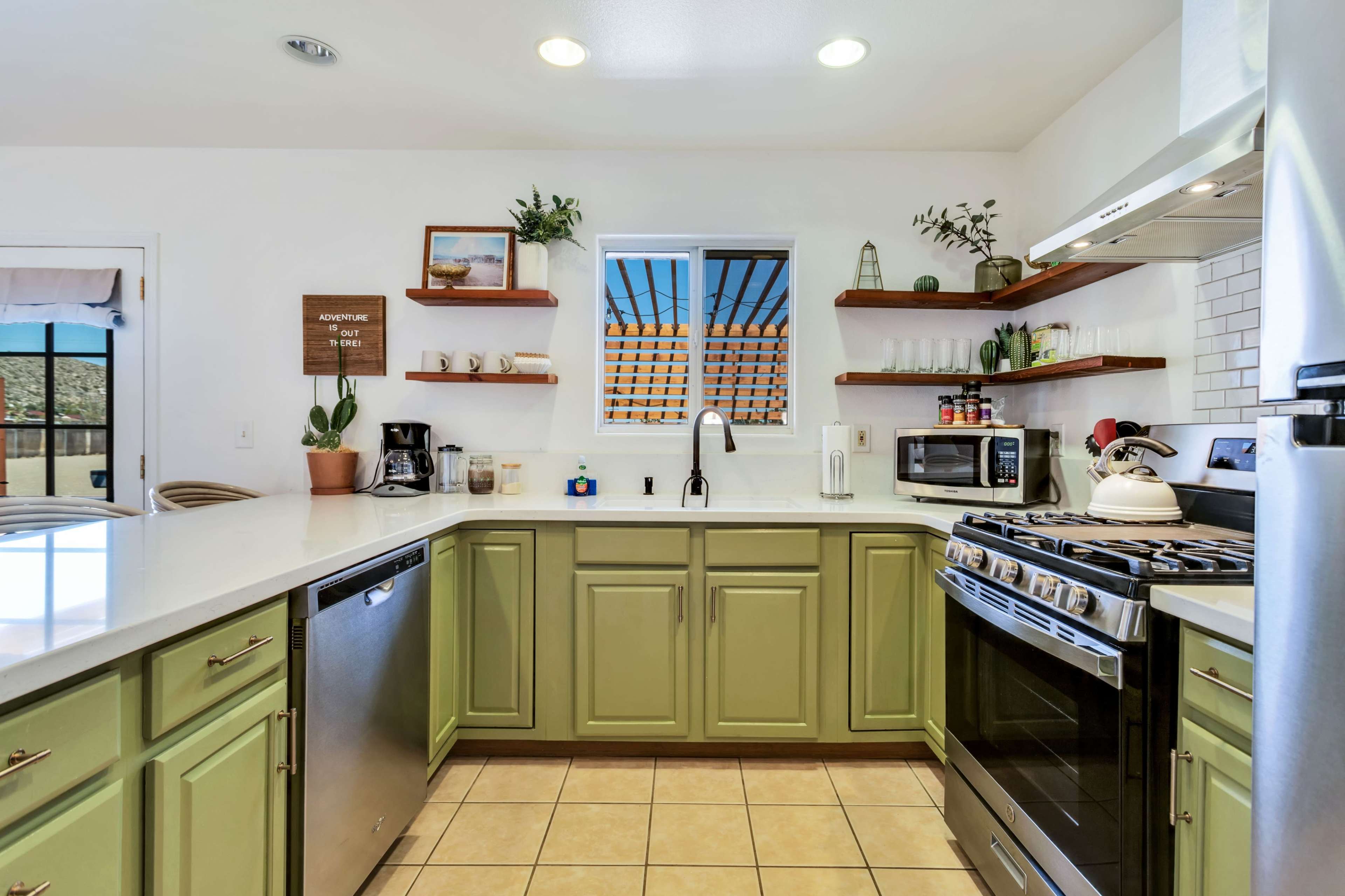 The kitchen features green cabinets, a white countertop, and stainless steel appliances with open shelving displaying plants and kitchenware.