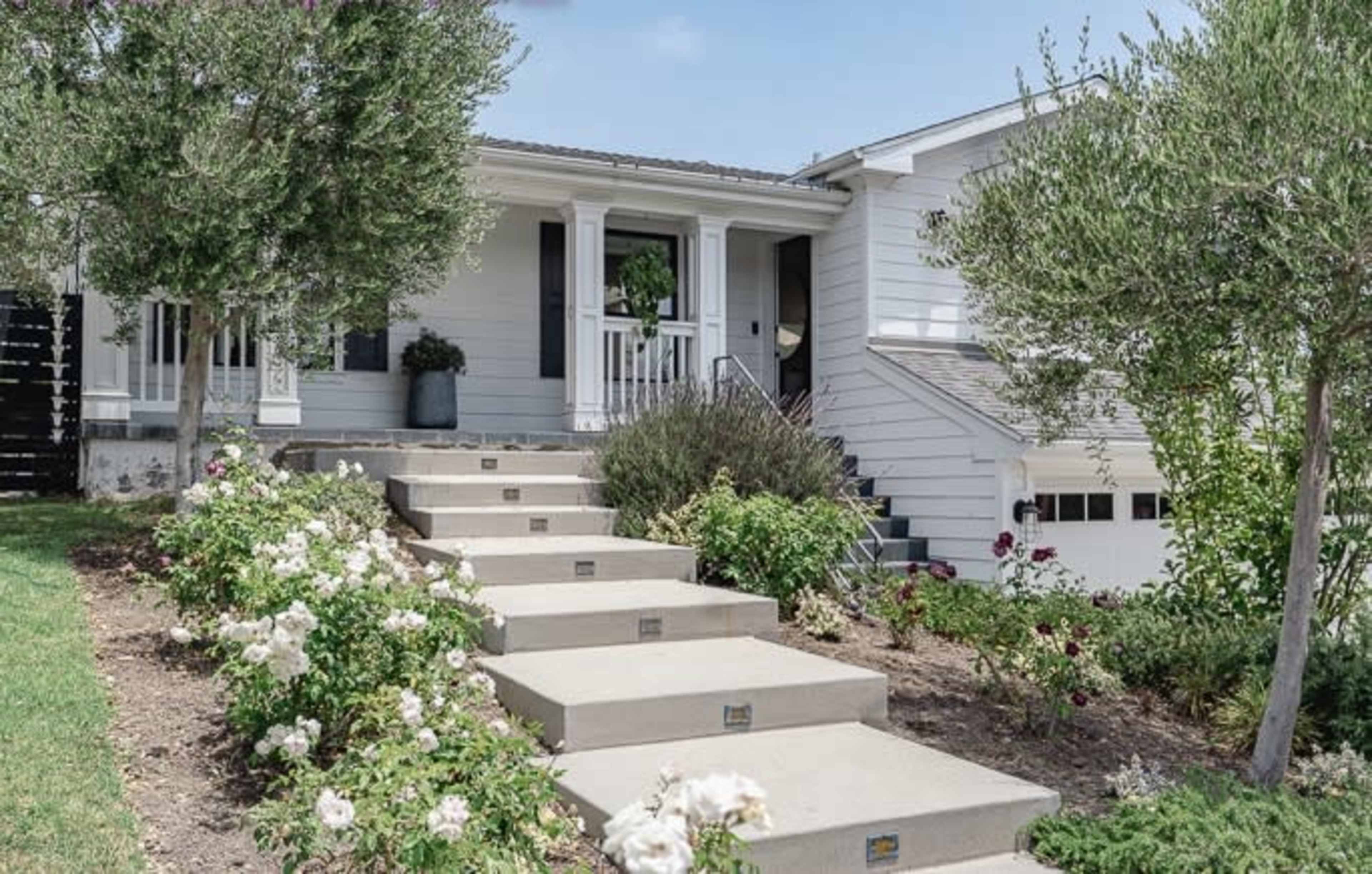 A house with a staircase leading up to a covered porch, surrounded by neatly landscaped flower beds and shrubs.