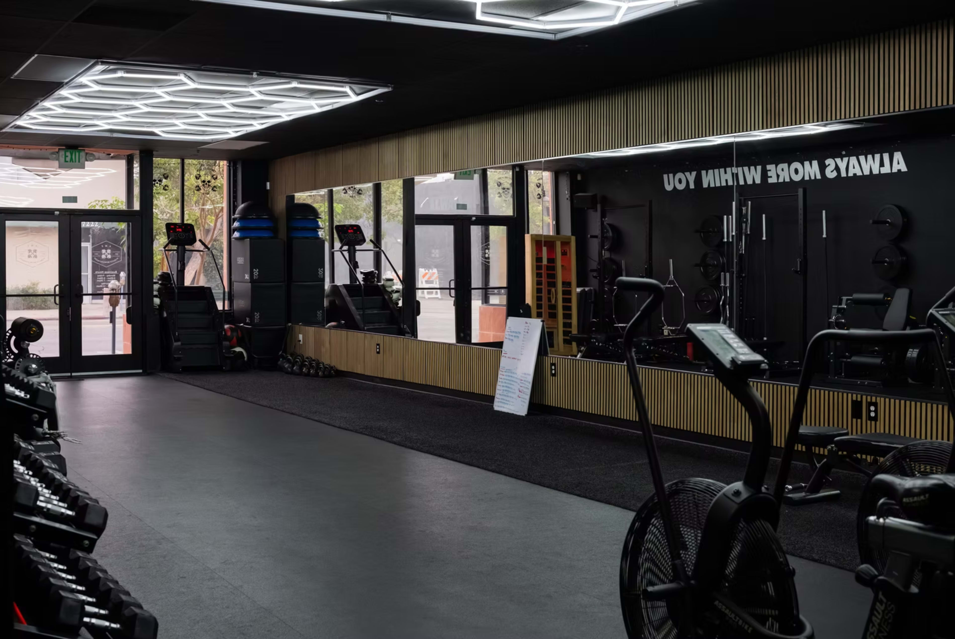 The image shows a modern gym interior with exercise equipment, including bikes and weights, arranged along mirrored walls and illuminated by overhead lighting.