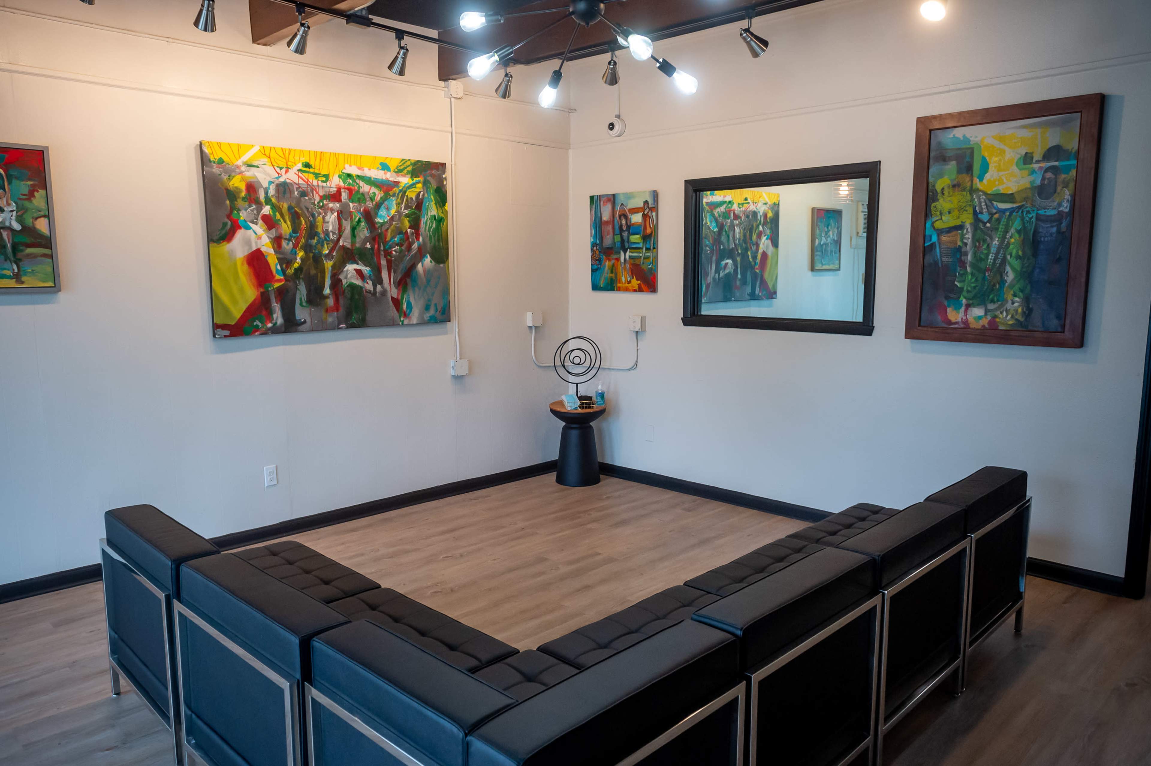 The image depicts an art gallery featuring colorful paintings on the walls and a seating arrangement in a U-shape in the center of the room.