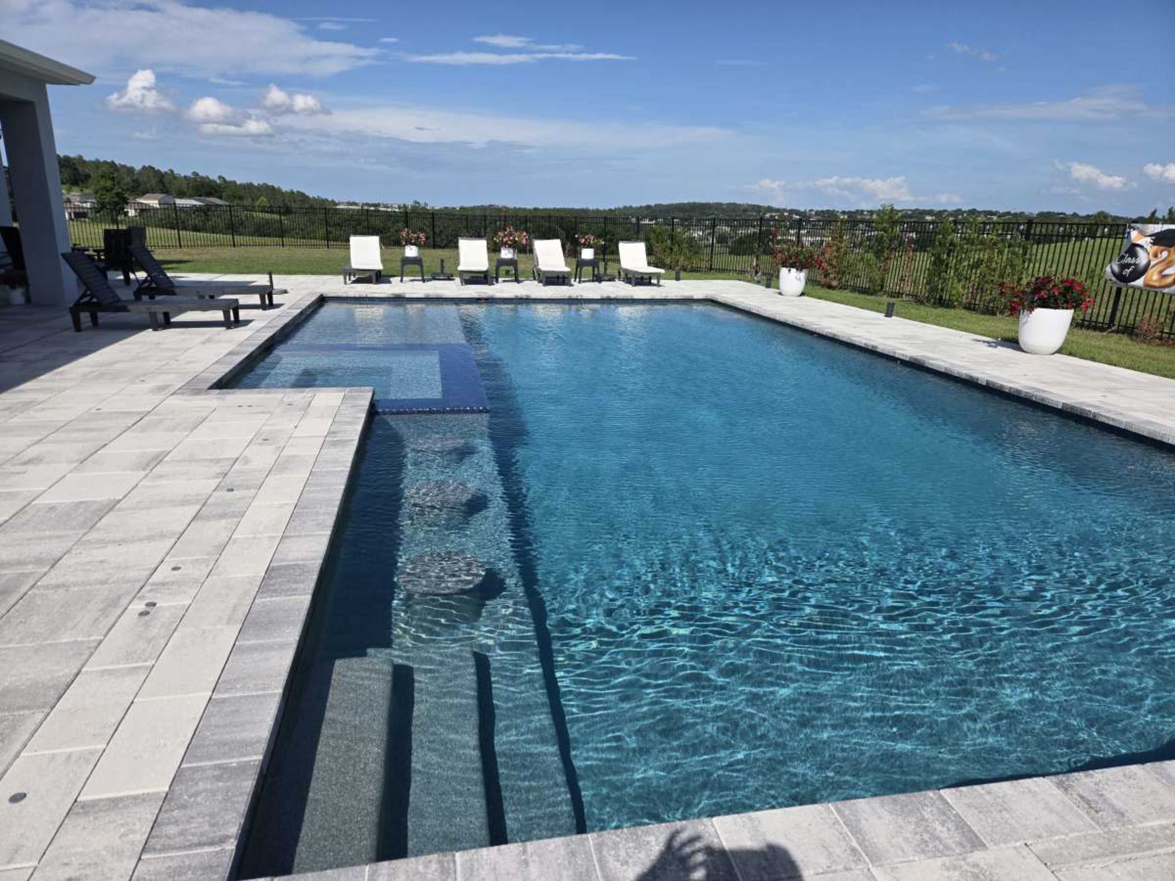 The image shows a blue swimming pool surrounded by lounge chairs and landscaped greenery under a clear sky.