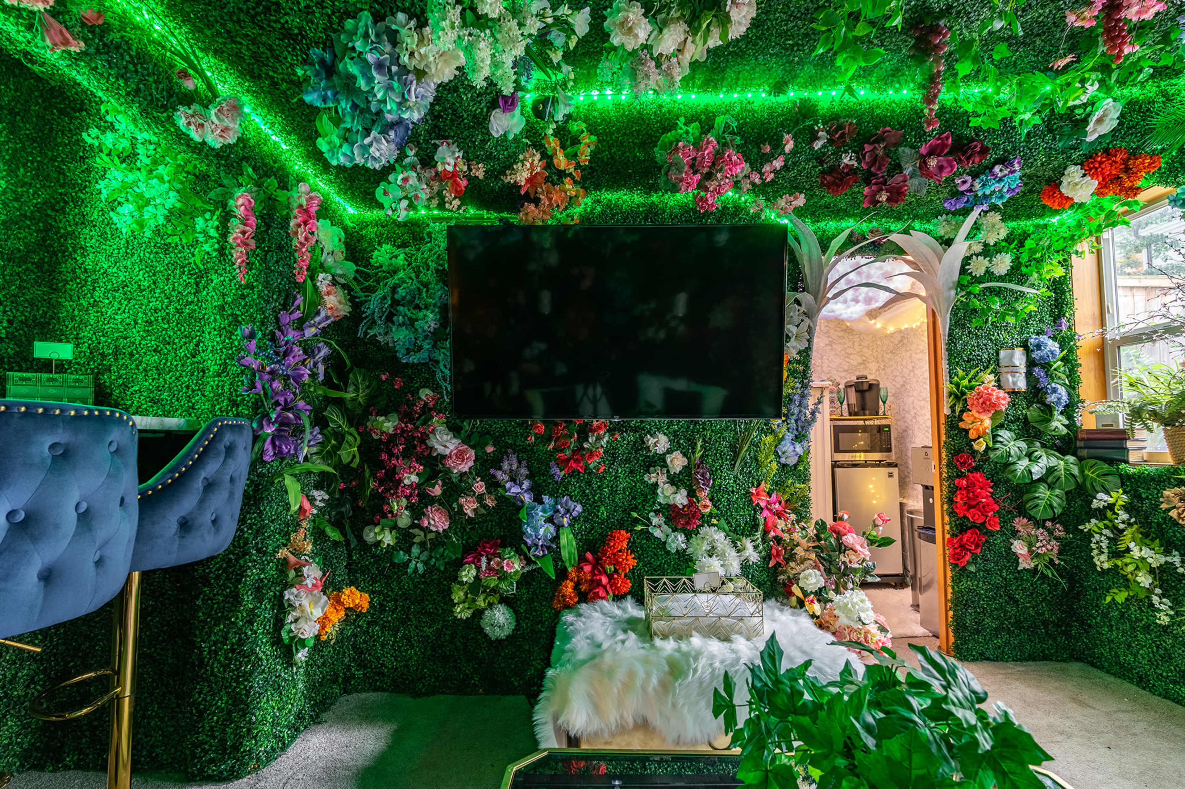 The room features walls covered in artificial greenery and colorful flowers, with a large television mounted on one wall and a doorway leading to a kitchen area.