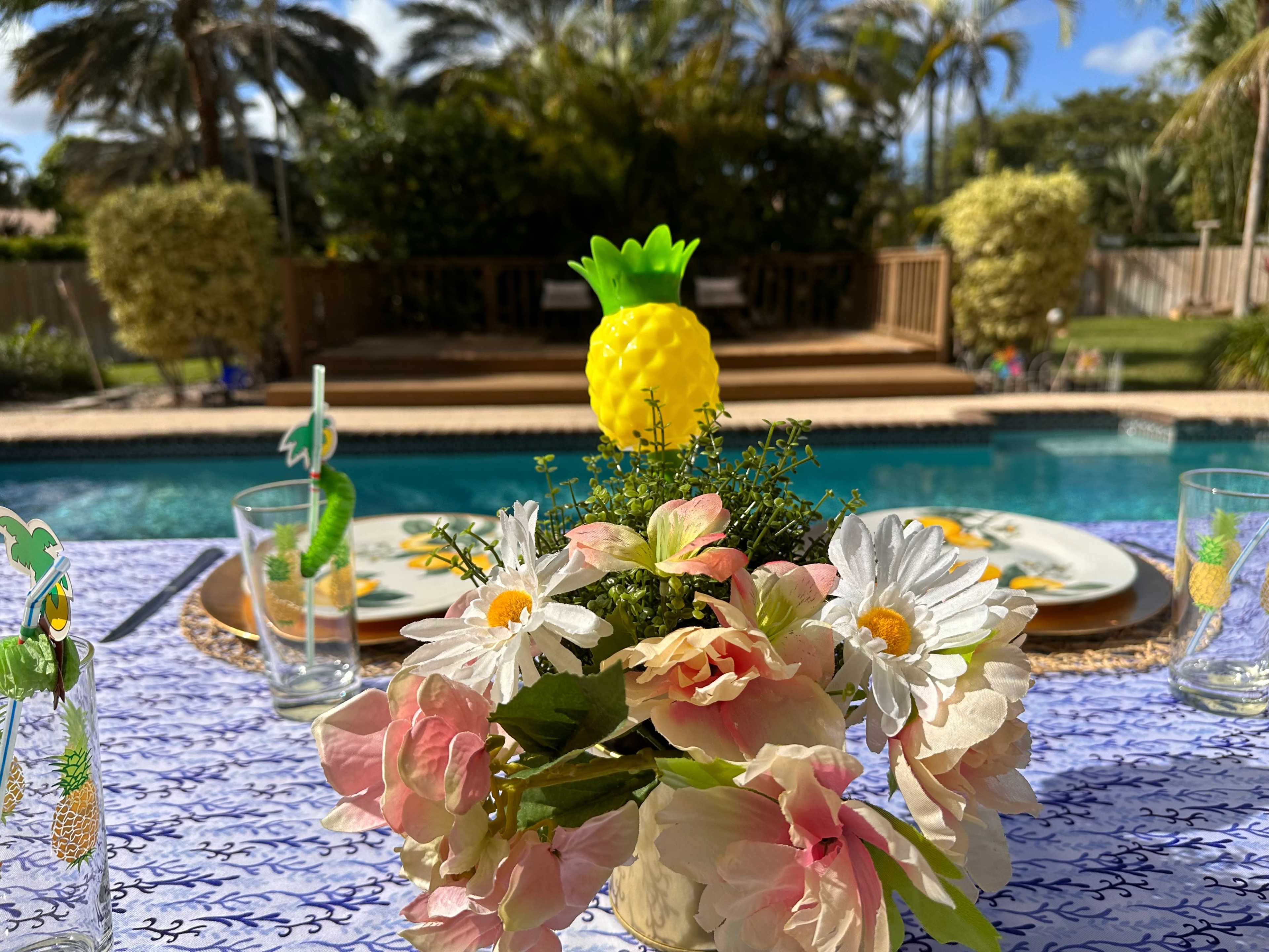 A table is set with a floral centerpiece and pineapple decoration, overlooking a swimming pool and palm trees in the background.