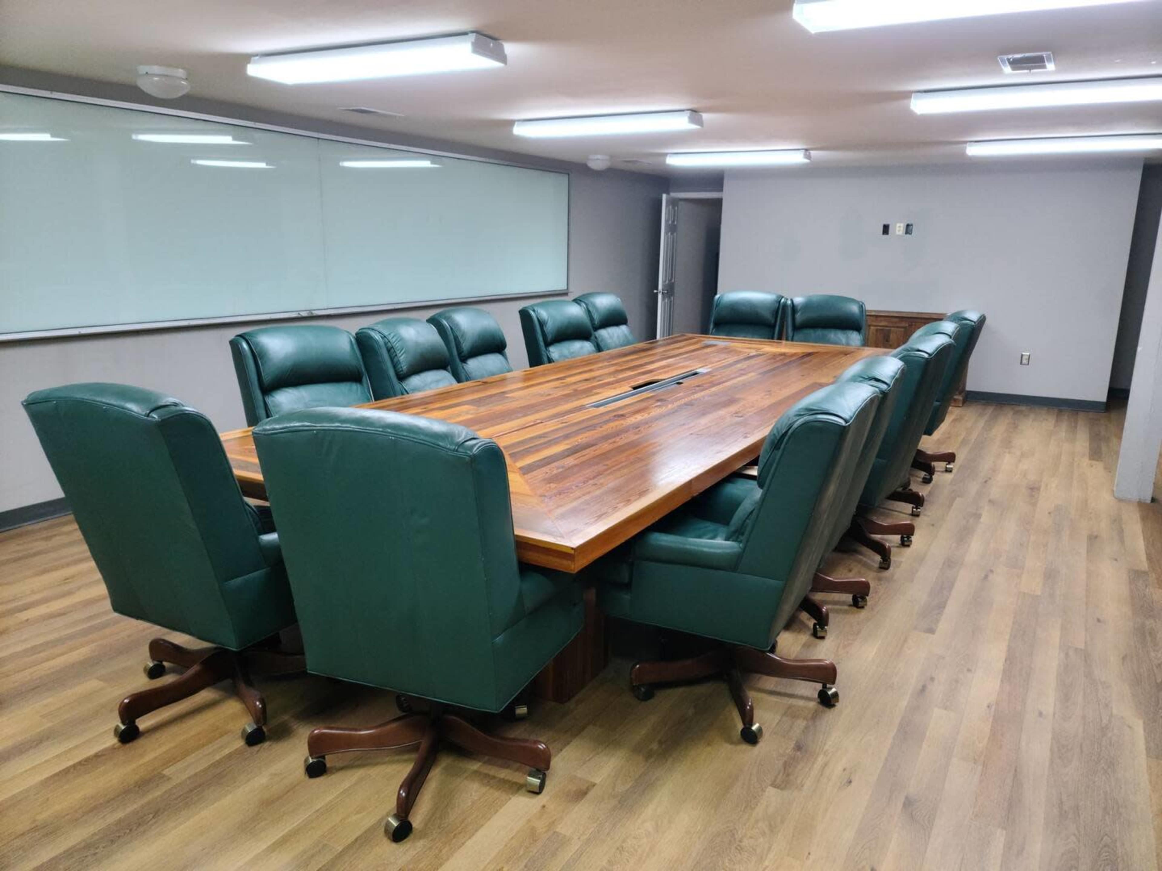 A conference room features a large wooden table surrounded by twelve green leather executive chairs on a hardwood floor.
