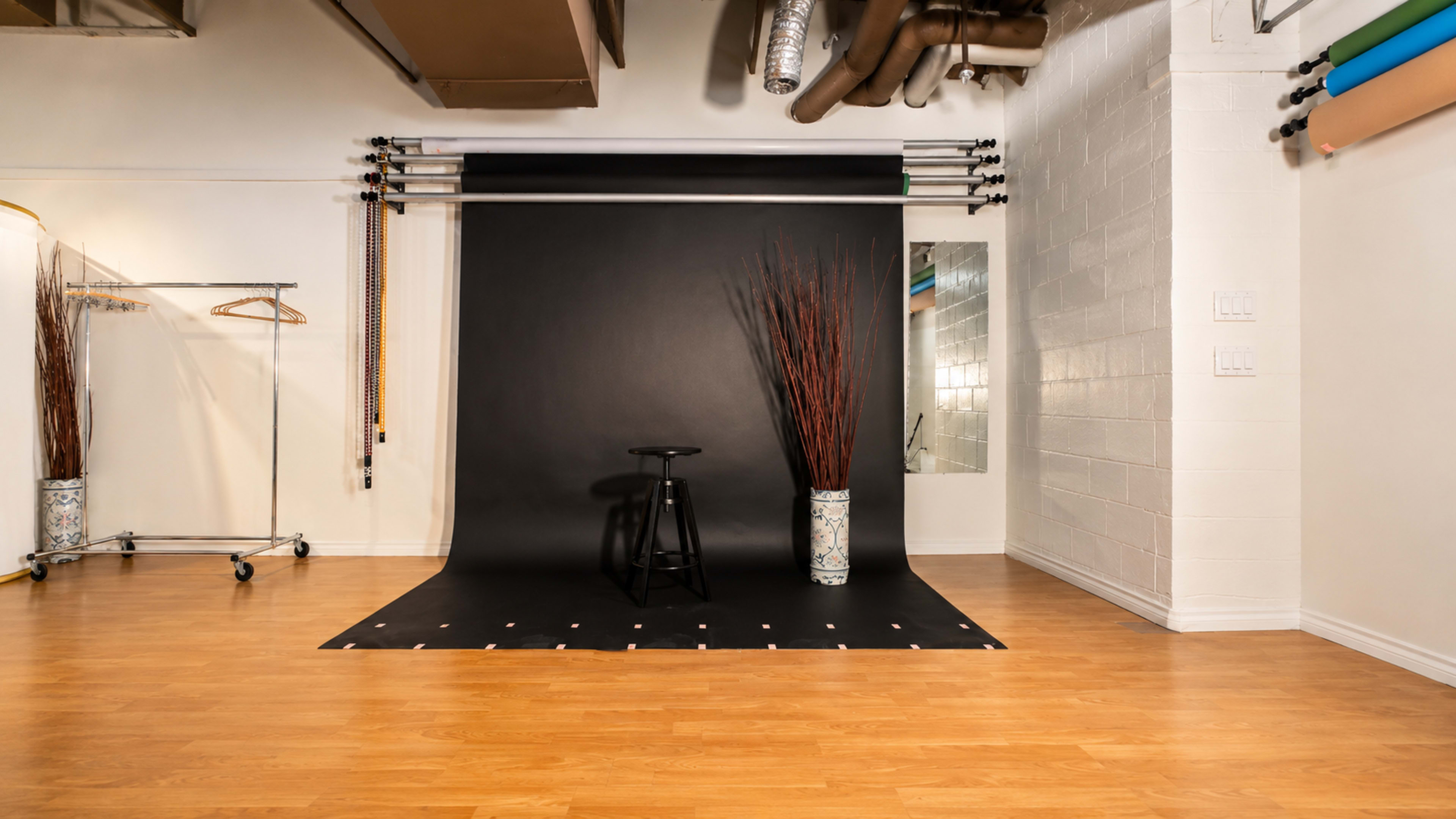 The image shows a photography studio featuring a black backdrop, a stool, two decorative vases with dry branches, and a clothing rack.