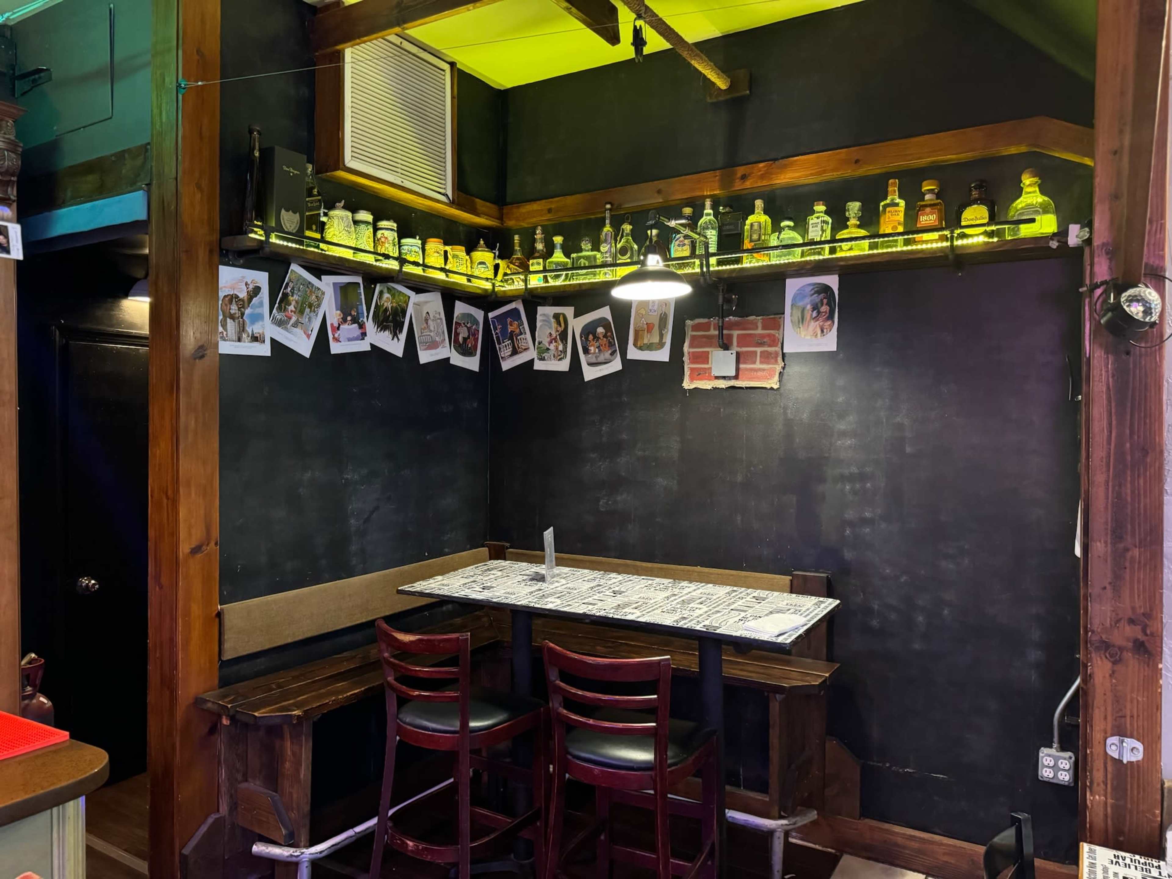 A small, dimly lit corner of a bar features a table with two wooden chairs and a shelf displaying various bottles above a wall adorned with black-and-white photos.