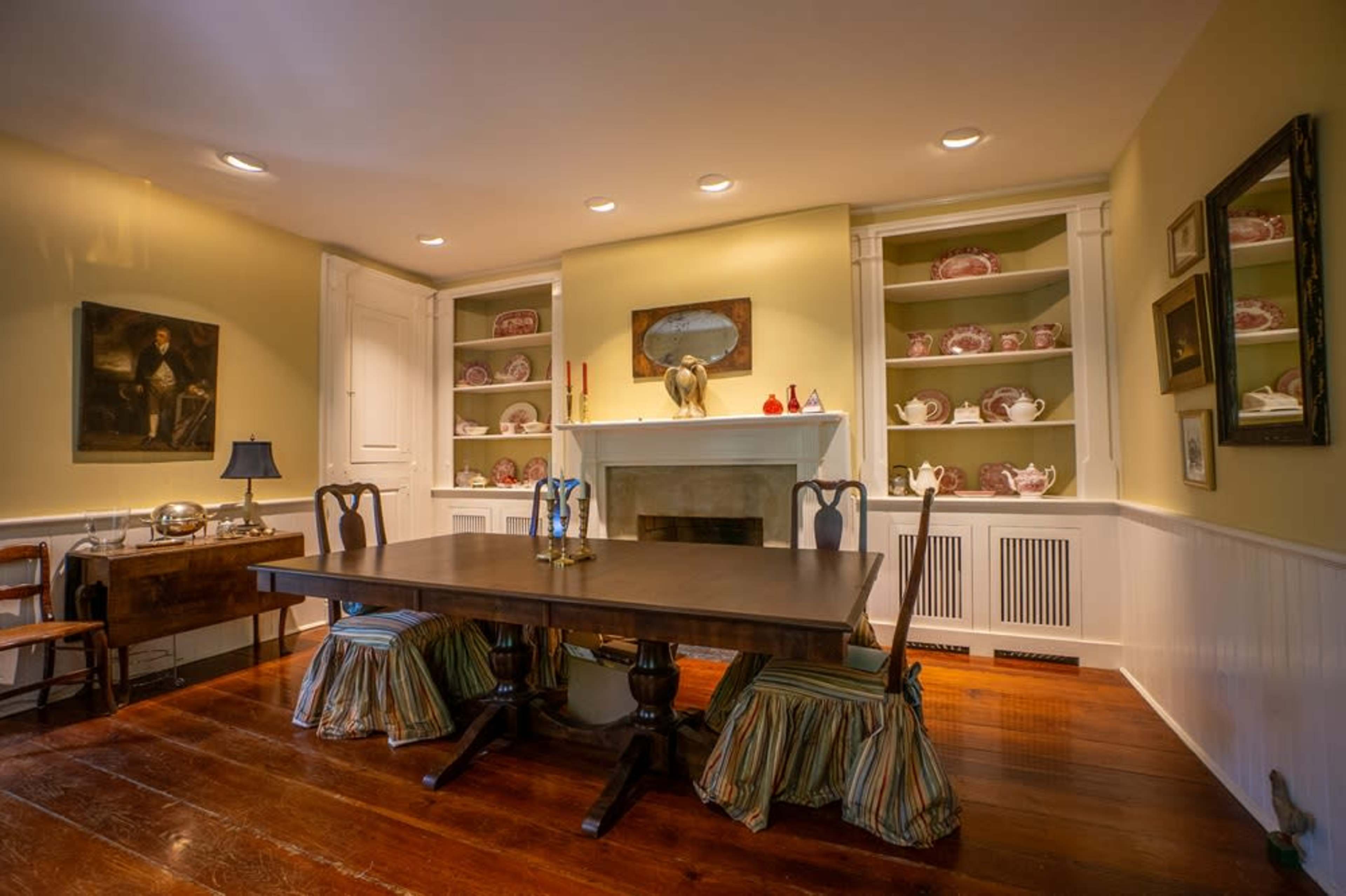A dining room with a wooden table surrounded by chairs, a decorative fireplace, and shelves filled with china.