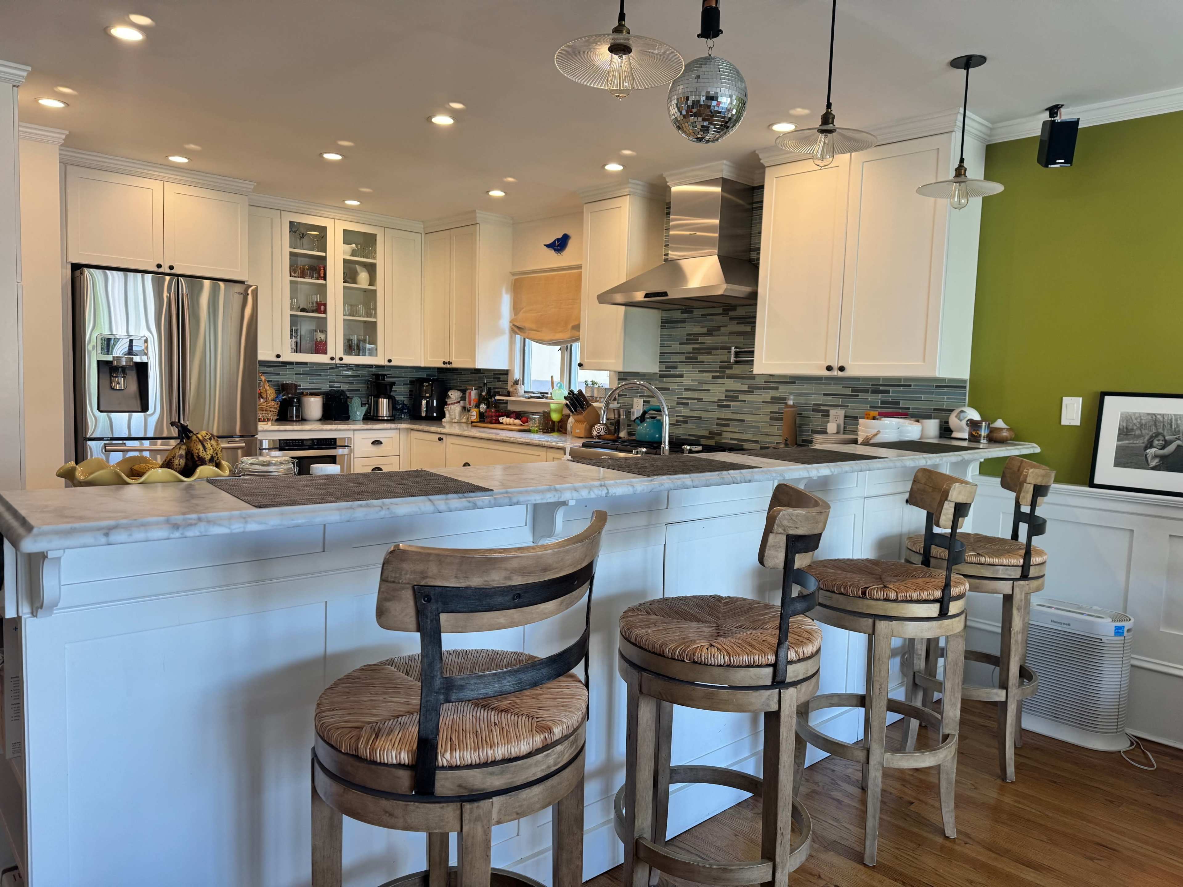 A modern kitchen features a central island with four wooden bar stools, stainless steel appliances, and green accent walls.
