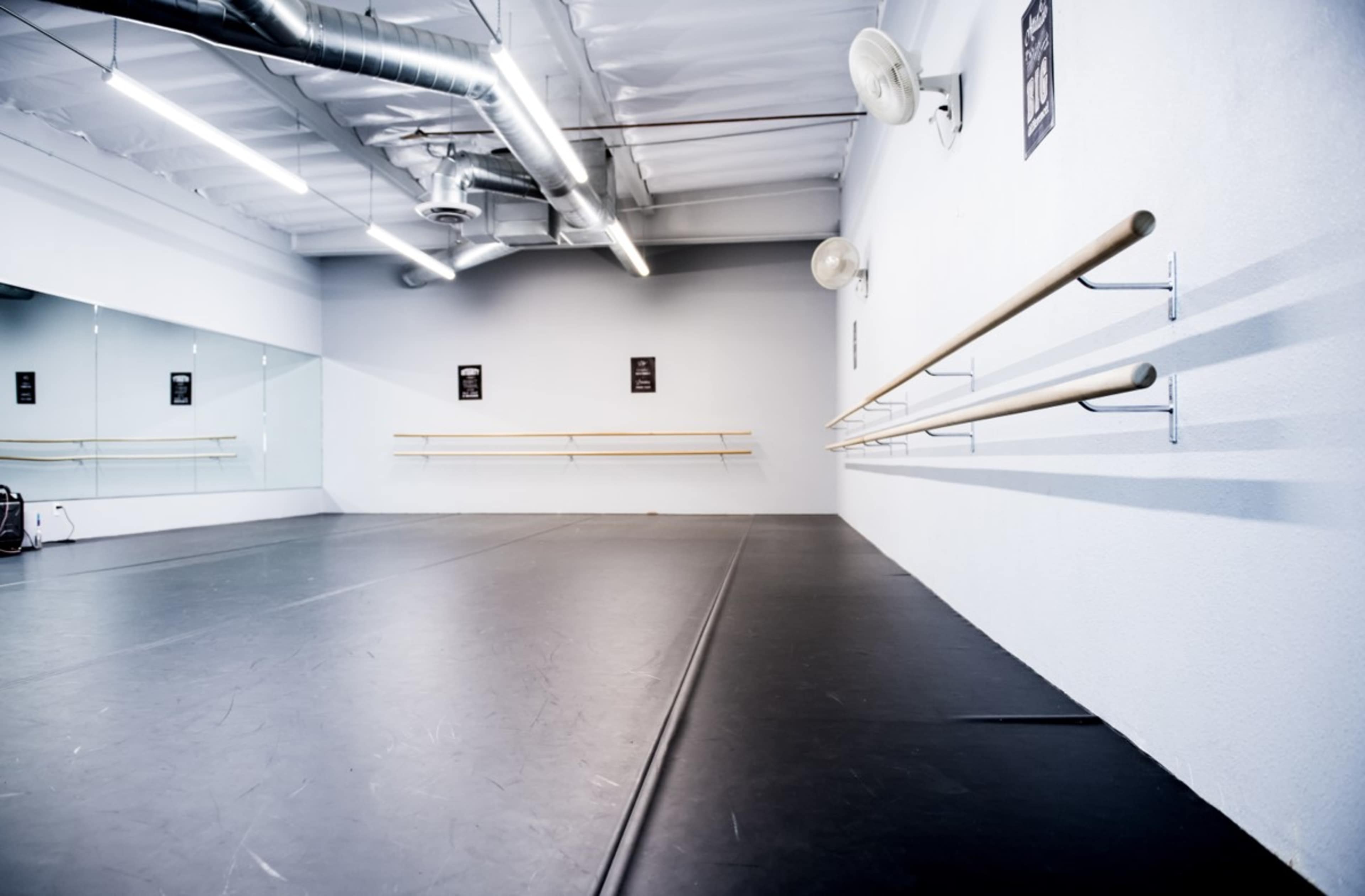 A spacious dance studio with mirrored walls, wooden ballet barres, and a black rubber floor.