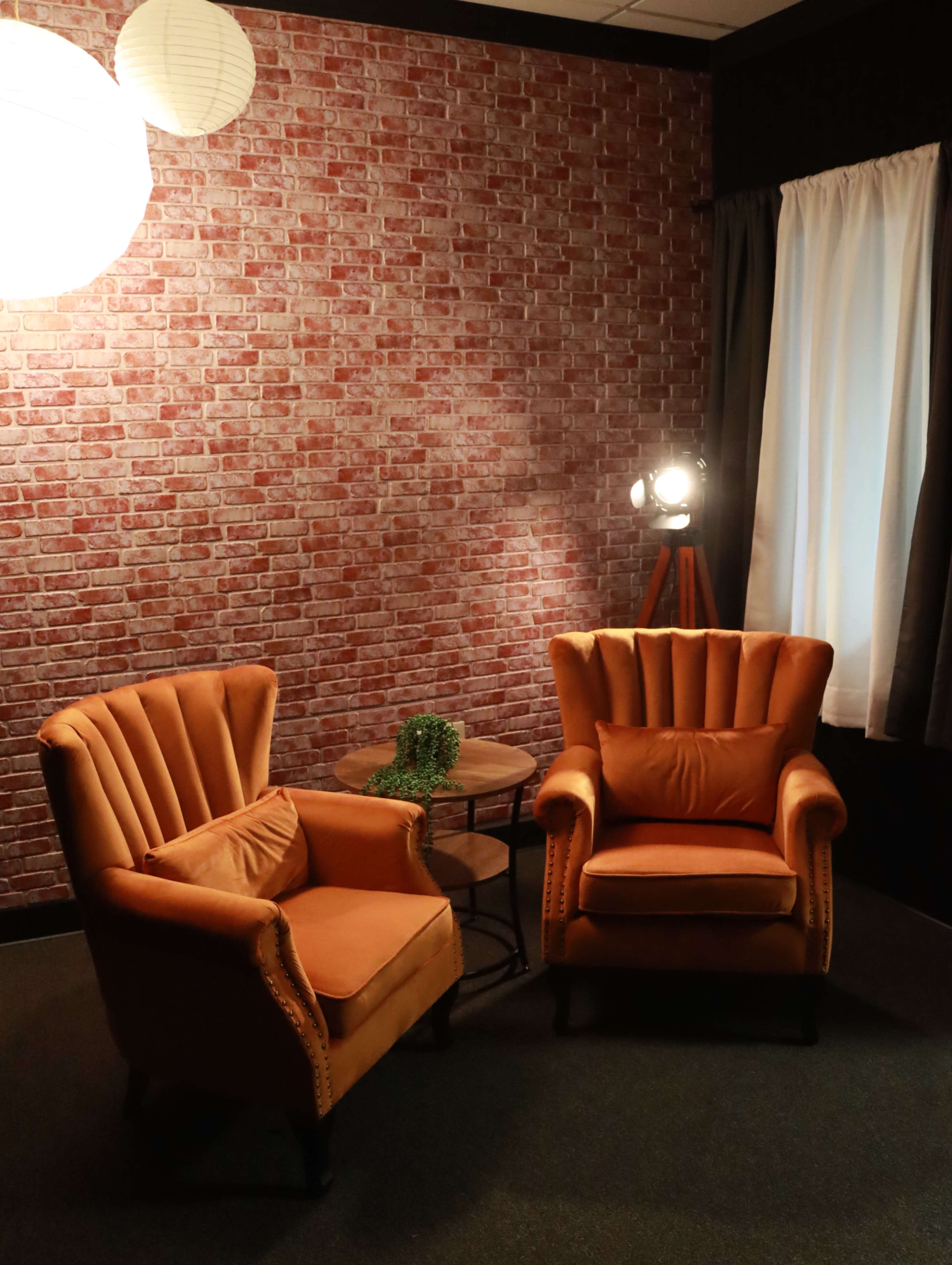 The image shows two orange armchairs arranged around a small wooden table in a room with a red brick wall and a lamp in the corner.