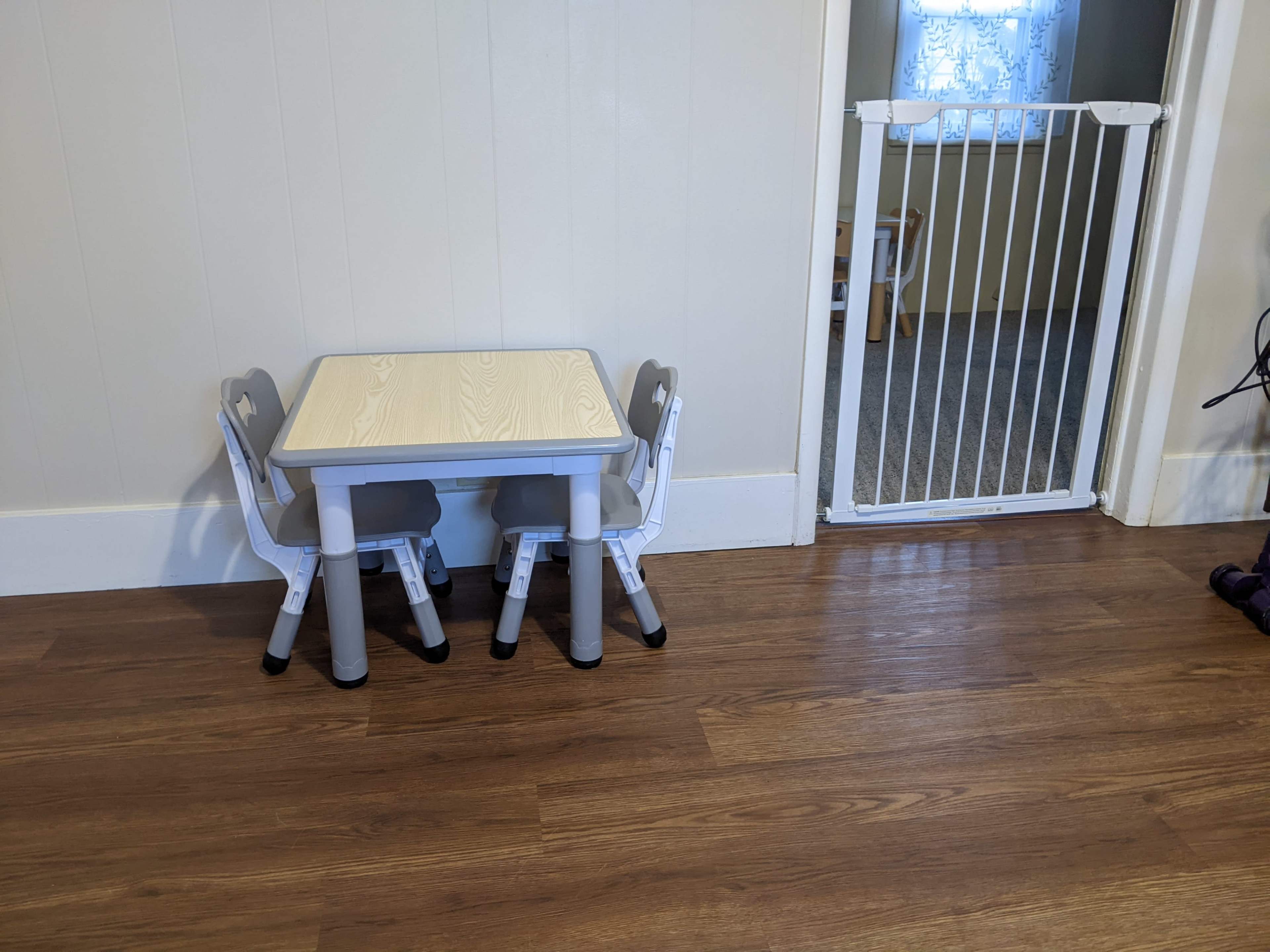 A small kids' table with two chairs is positioned against a wall next to a baby gate in a room with wooden flooring.