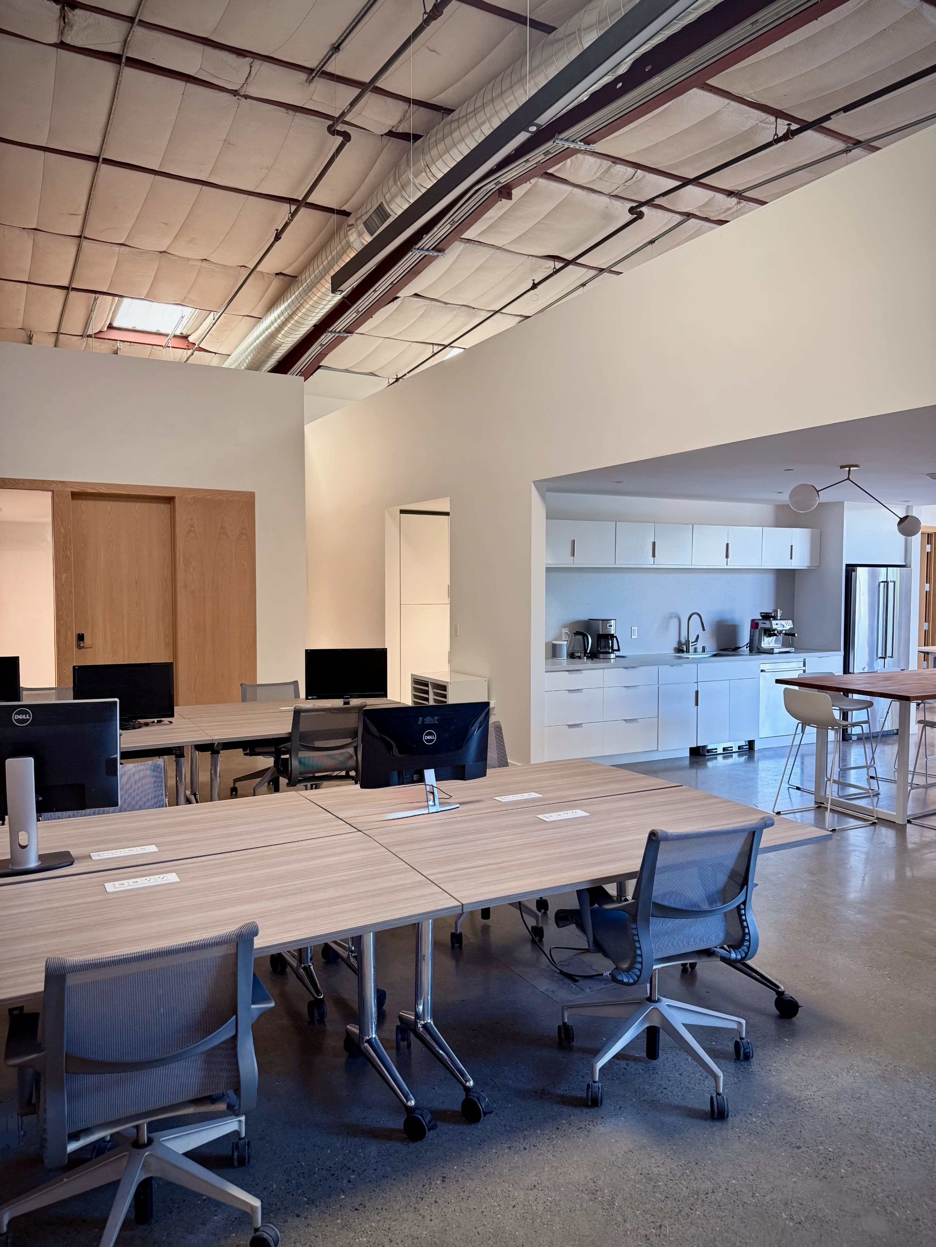 The image shows a modern office space with multiple desks and a kitchenette area in the background.
