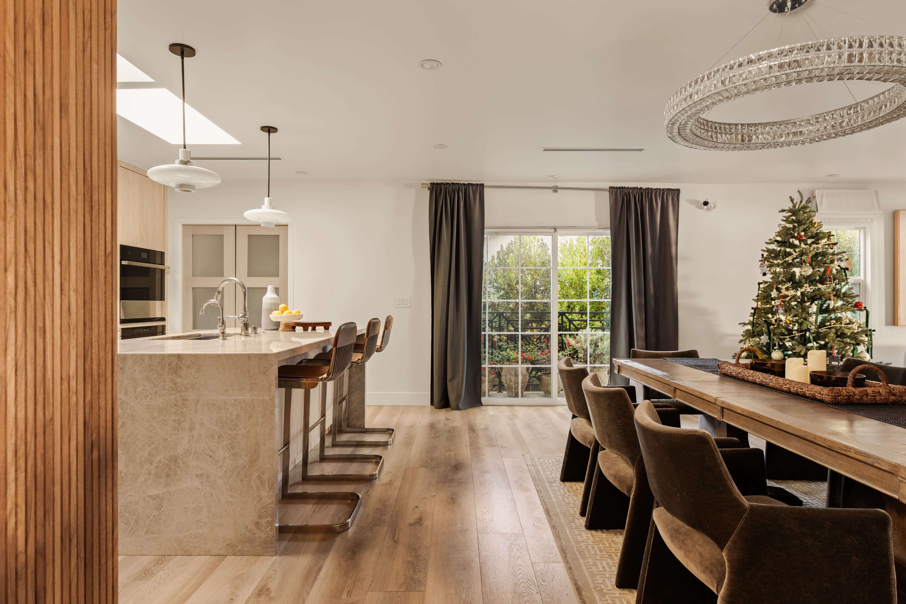A modern kitchen and dining area feature a large wooden table, a beautifully decorated Christmas tree, and stylish bar stools along a marble countertop.