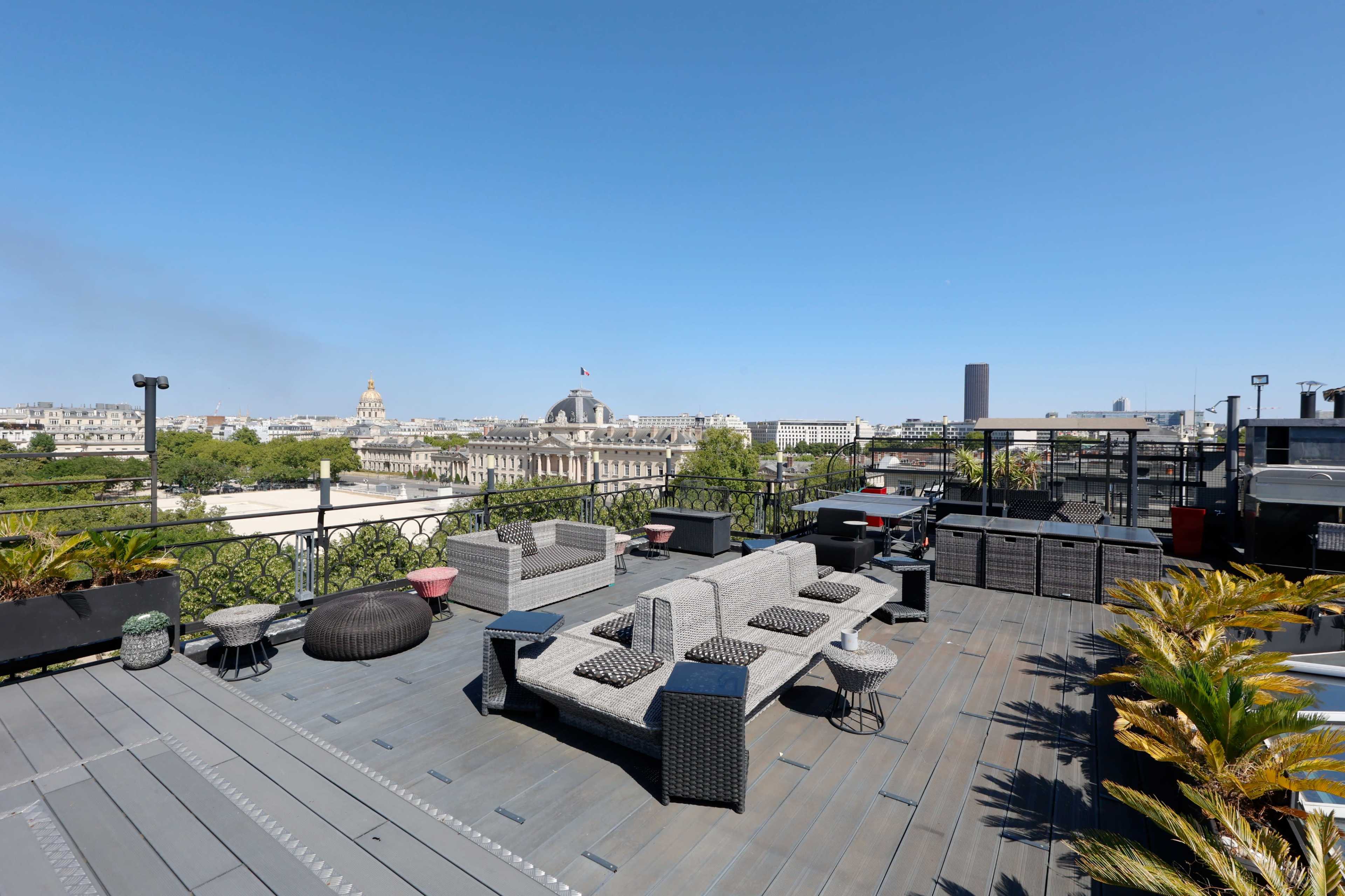 The image shows a spacious rooftop terrace adorned with outdoor furniture and offering a view of distant buildings and clear blue skies.