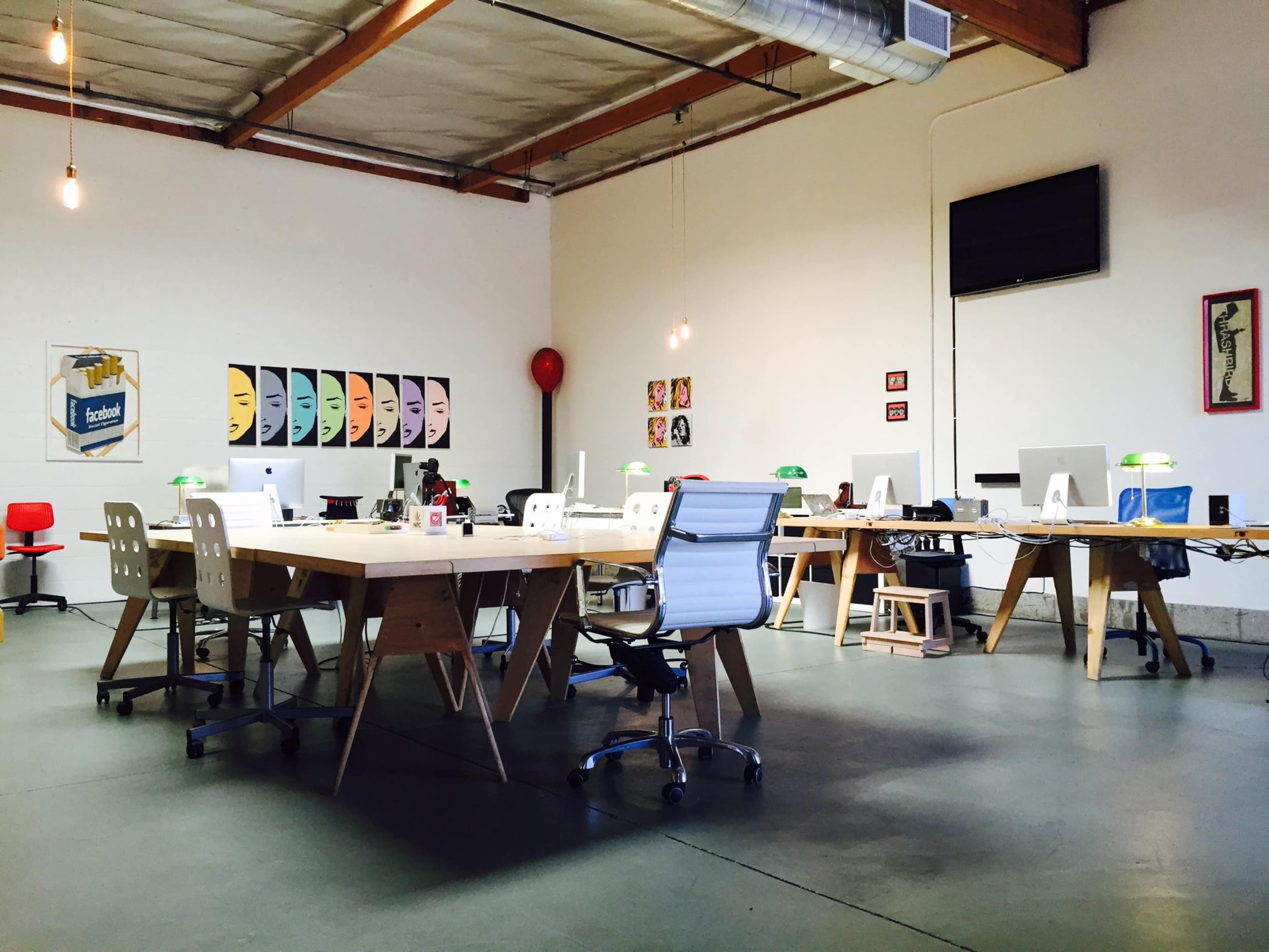 The image shows a modern office space with wooden desks, stylish chairs, and various decorative items against white walls.