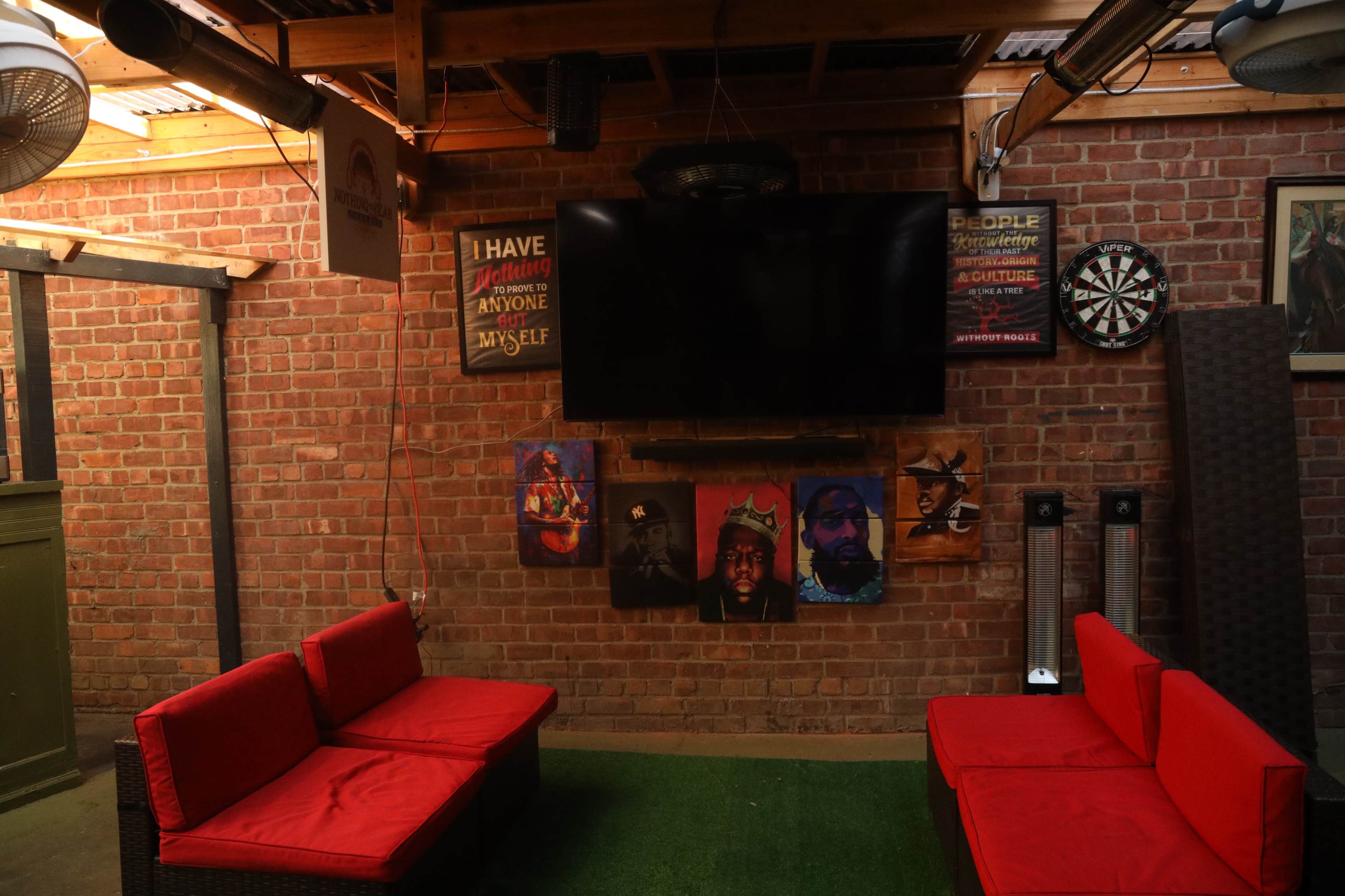 A cozy indoor space with red seating, a large TV mounted on a brick wall, and artwork featuring portraits on display.