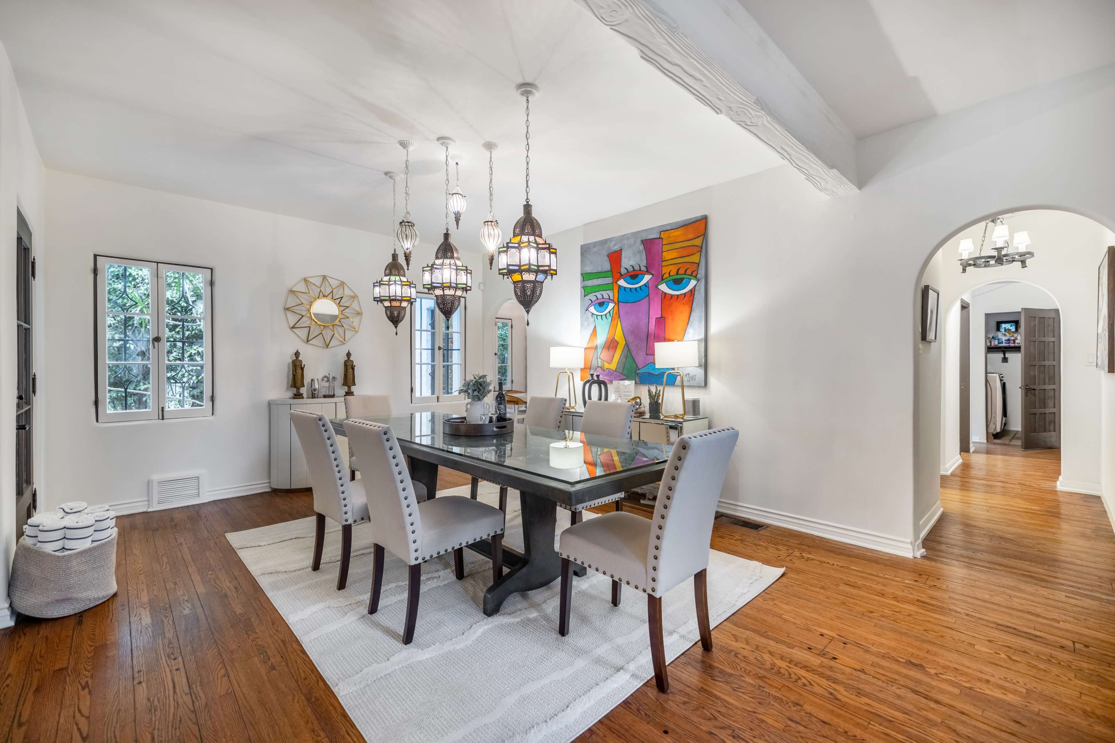 A dining room features a rectangular table surrounded by upholstered chairs, with pendant lights hanging above and colorful artwork on the wall.