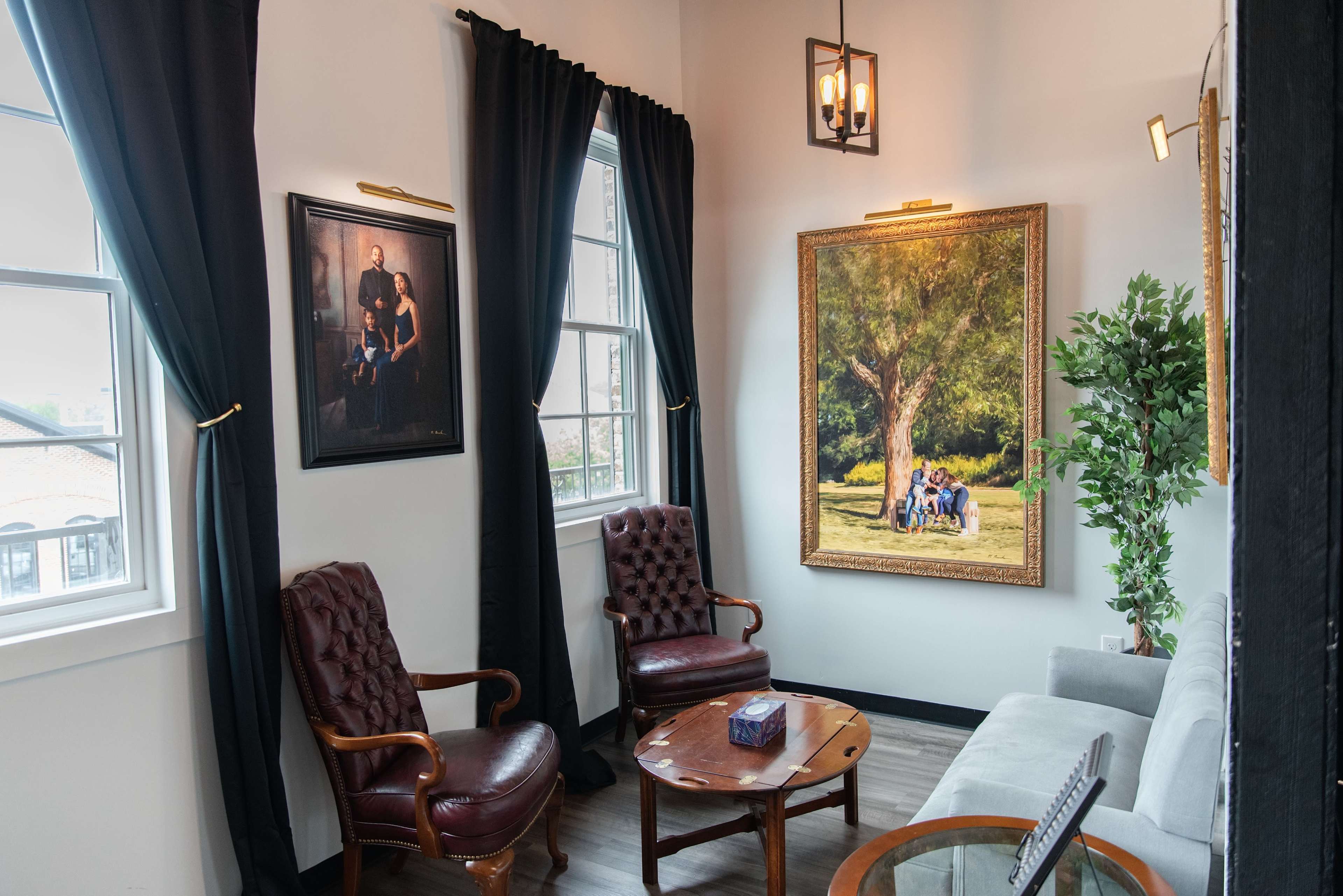 The image shows a corner of a room featuring two armchairs, a small round table, and framed artwork on the walls, with large windows allowing natural light in.