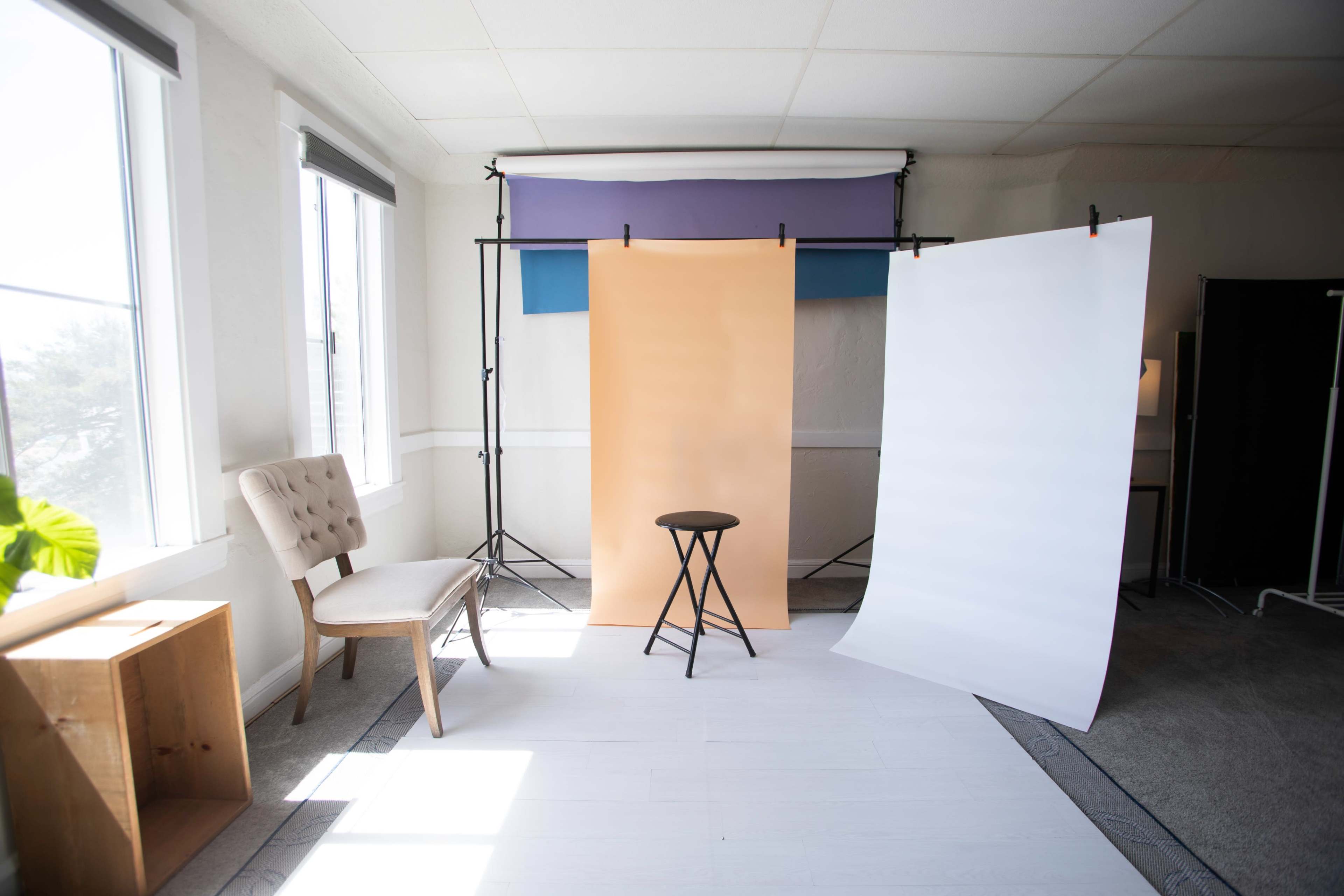 A photography studio setup with a stool in front of three backdrops: one orange, one white, and one purple, along with a chair and natural light coming through the windows.