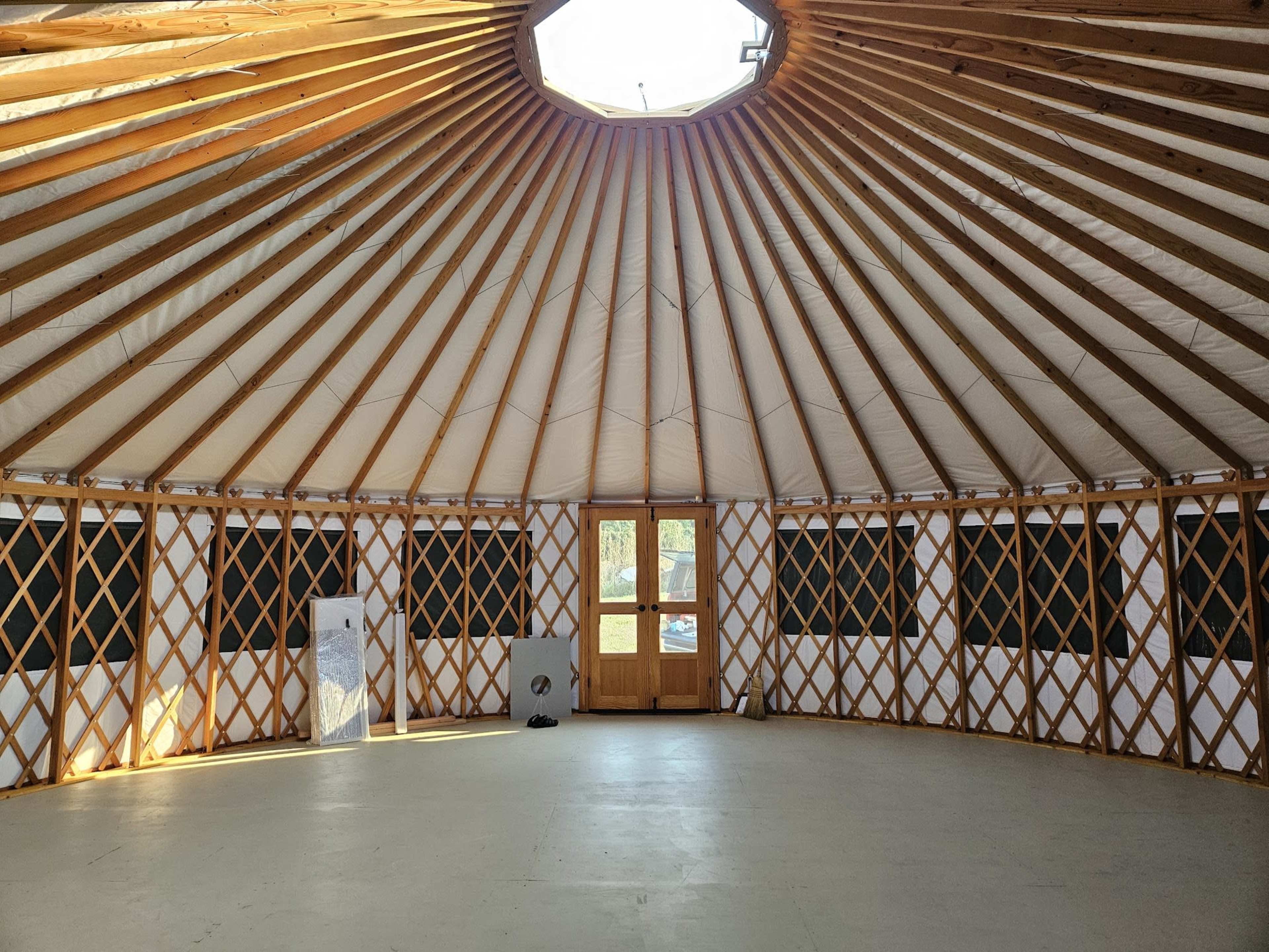 The interior of a yurt features wooden beams radiating from a central circular opening, with a wooden door and windows along the walls.