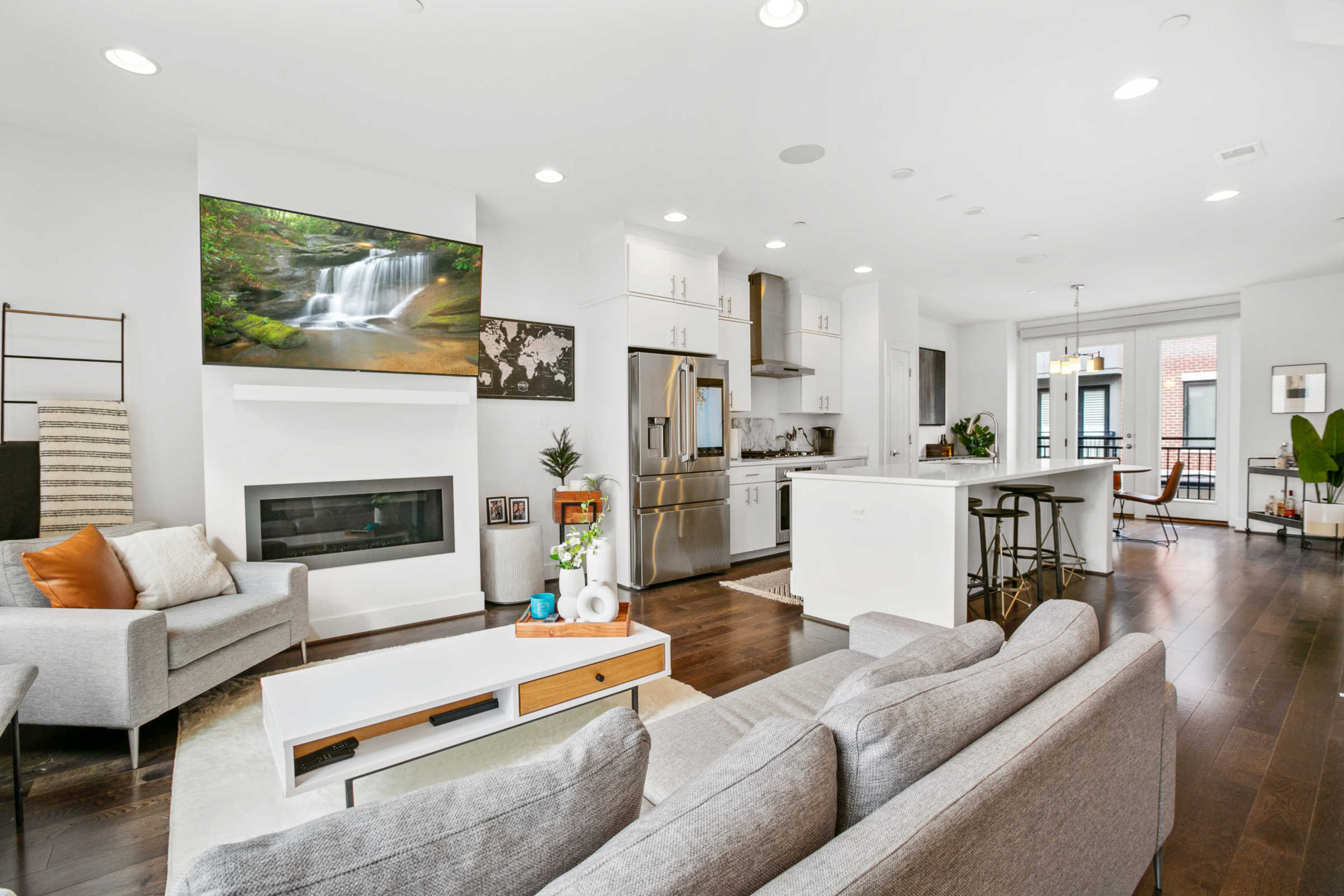 A modern living room and kitchen area featuring a large television mounted on the wall, a sofa, a dining table, and a balcony view.