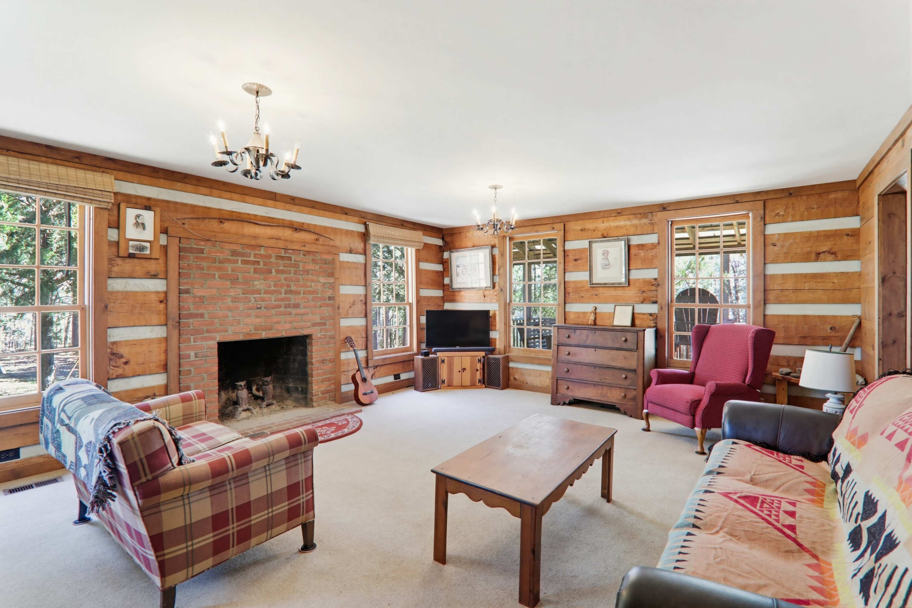 The image shows a living room with wooden walls, a brick fireplace, a television, and a mix of chairs and a sofa arranged around a coffee table.