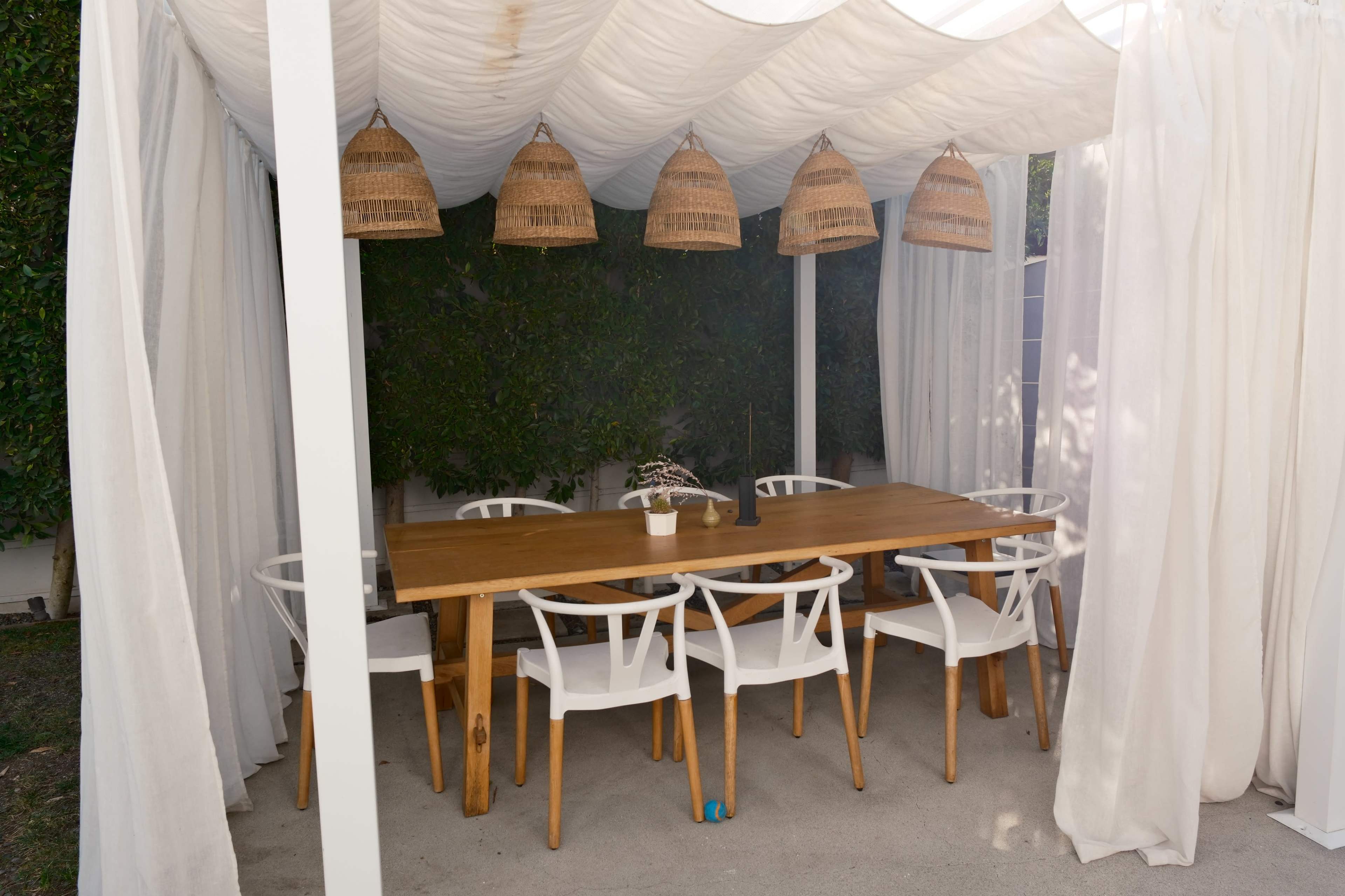A wooden table surrounded by white chairs is set under a canopy with hanging woven lamps, enclosed by sheer curtains and greenery.