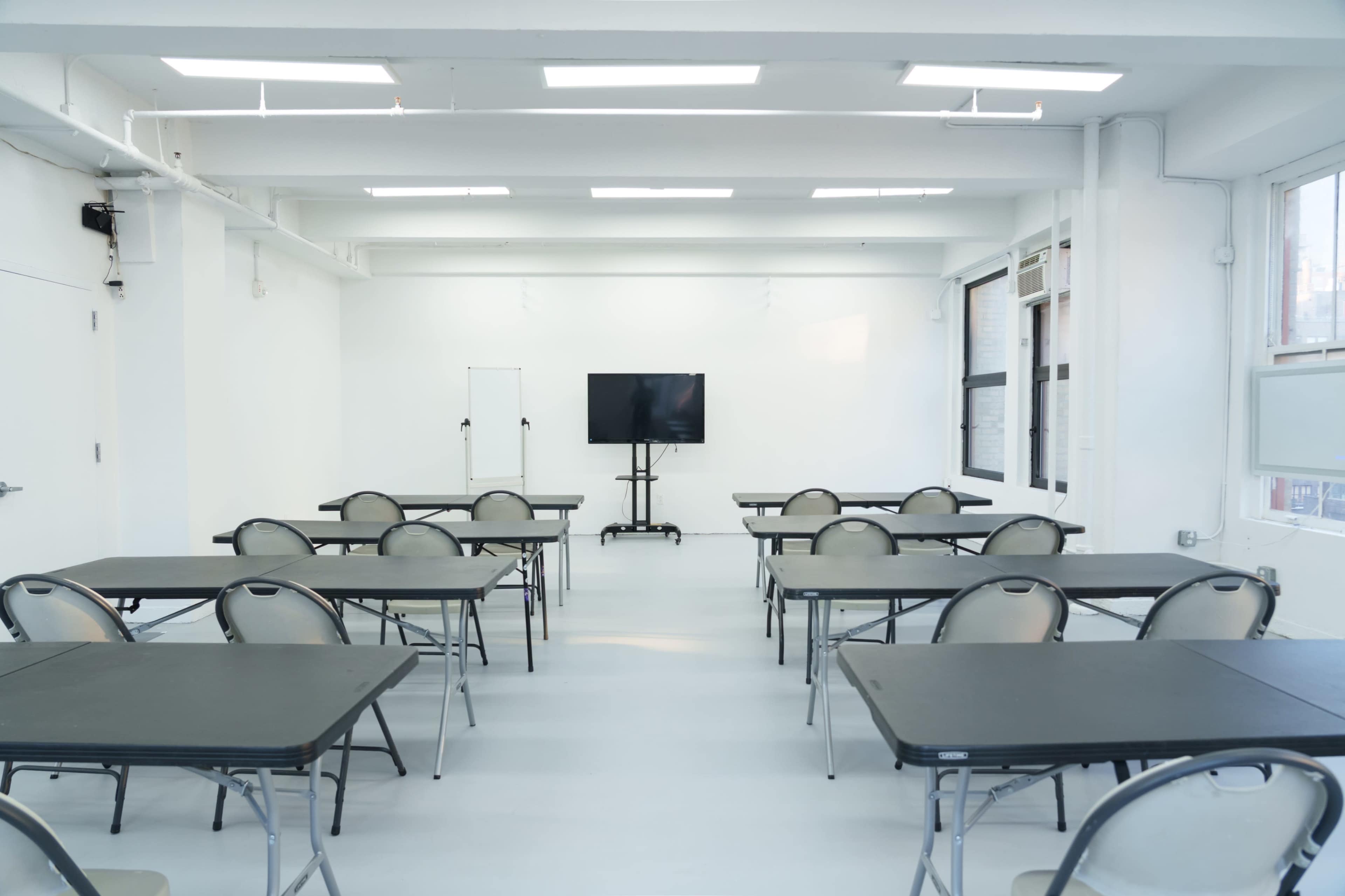 The image shows a spacious, empty classroom with rows of tables and chairs arranged around a central area, featuring a large screen mounted on a stand at the front.