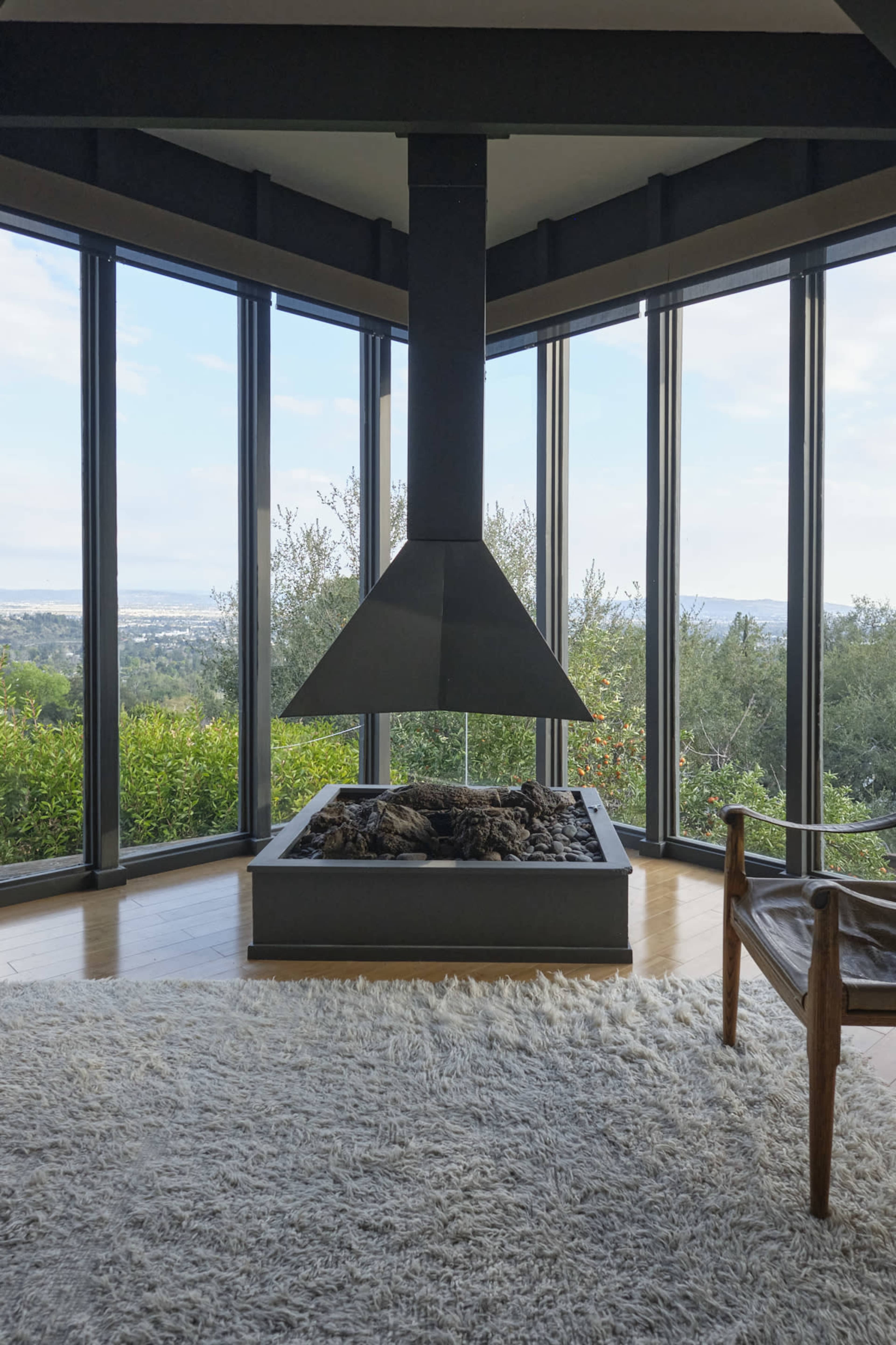 A modern, suspended fireplace is positioned in a spacious room with large glass windows offering a view of greenery and distant hills.