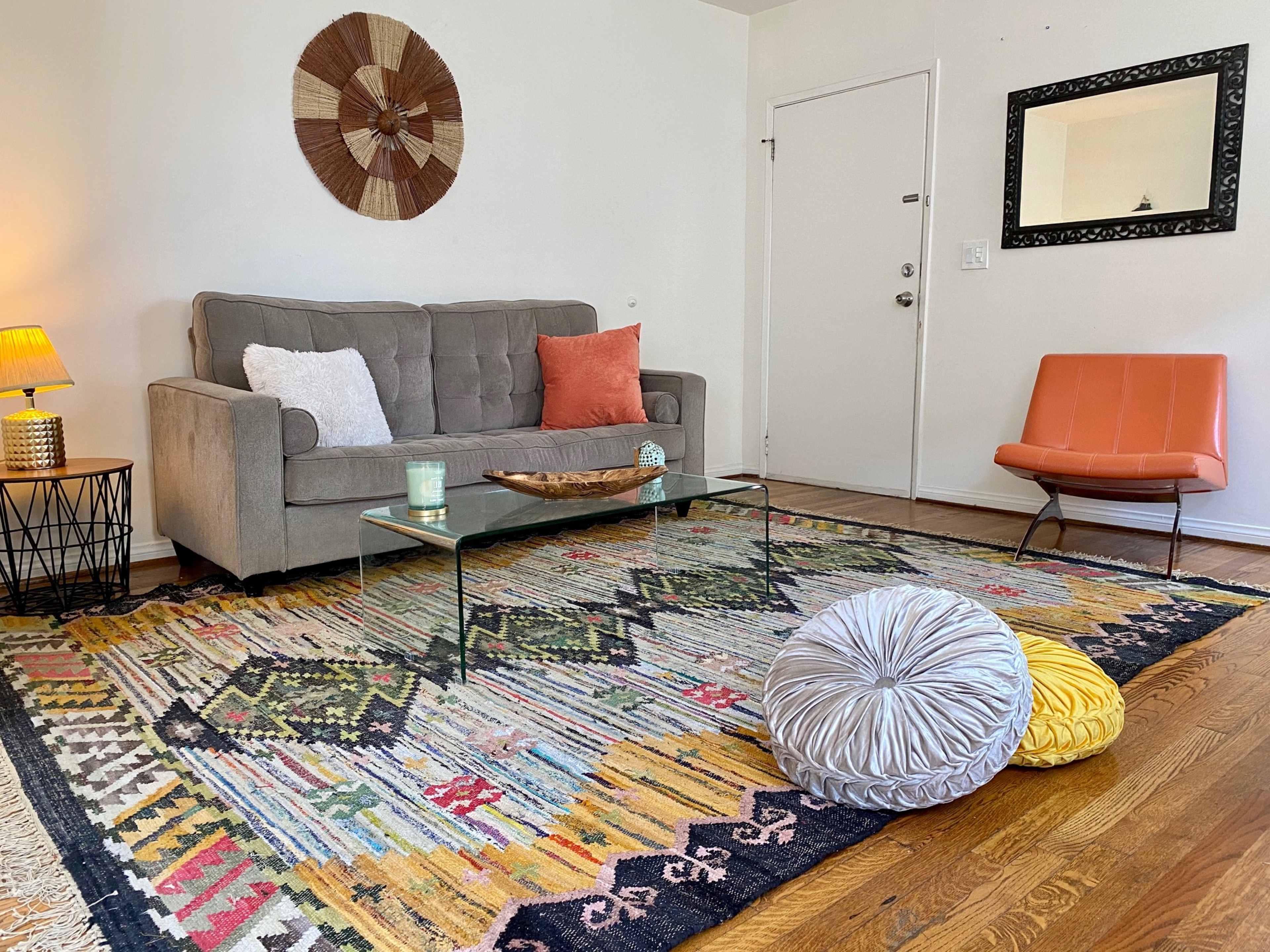 The scene shows a cozy living room with a grey sofa, a glass coffee table, a patterned area rug, a decorative wall hanging, and an orange accent chair.