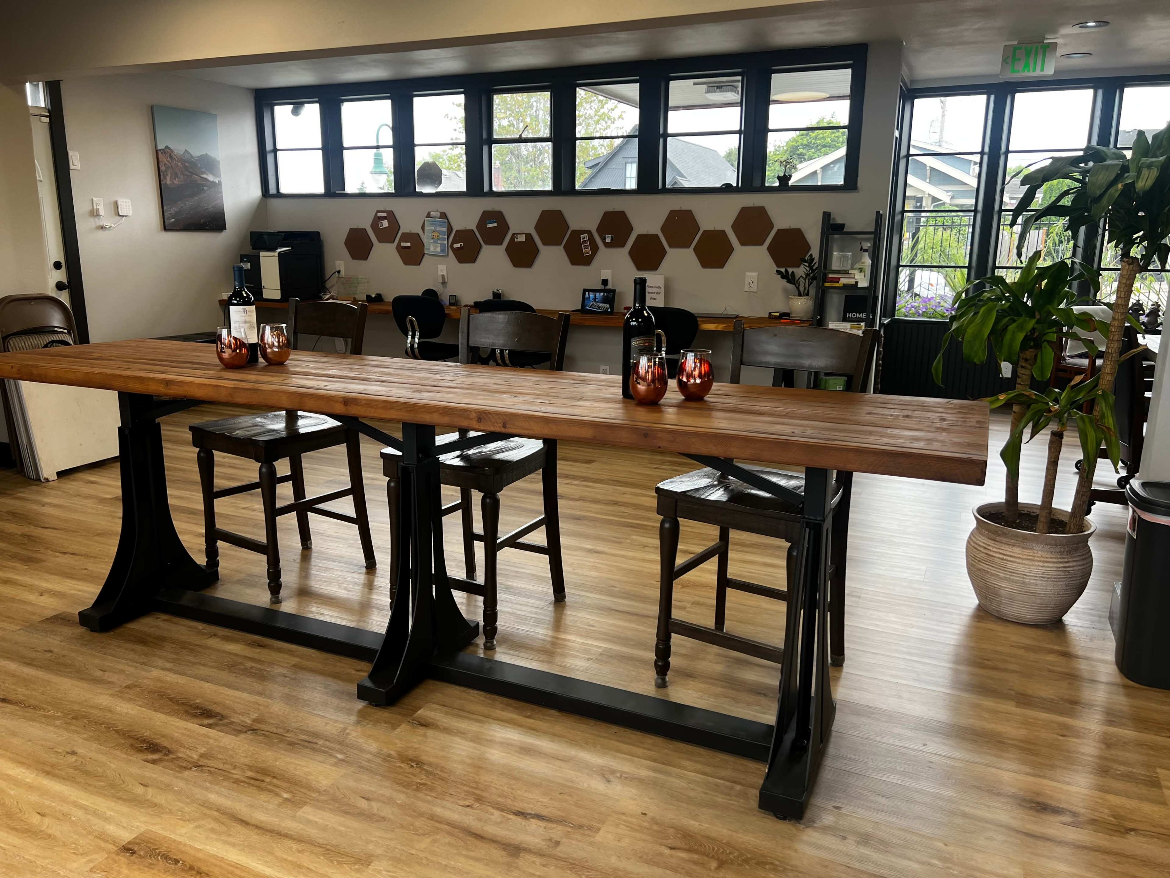 The image shows a spacious café interior with a wooden table surrounded by high chairs, shelves displaying plants, and hexagonal wall decor.