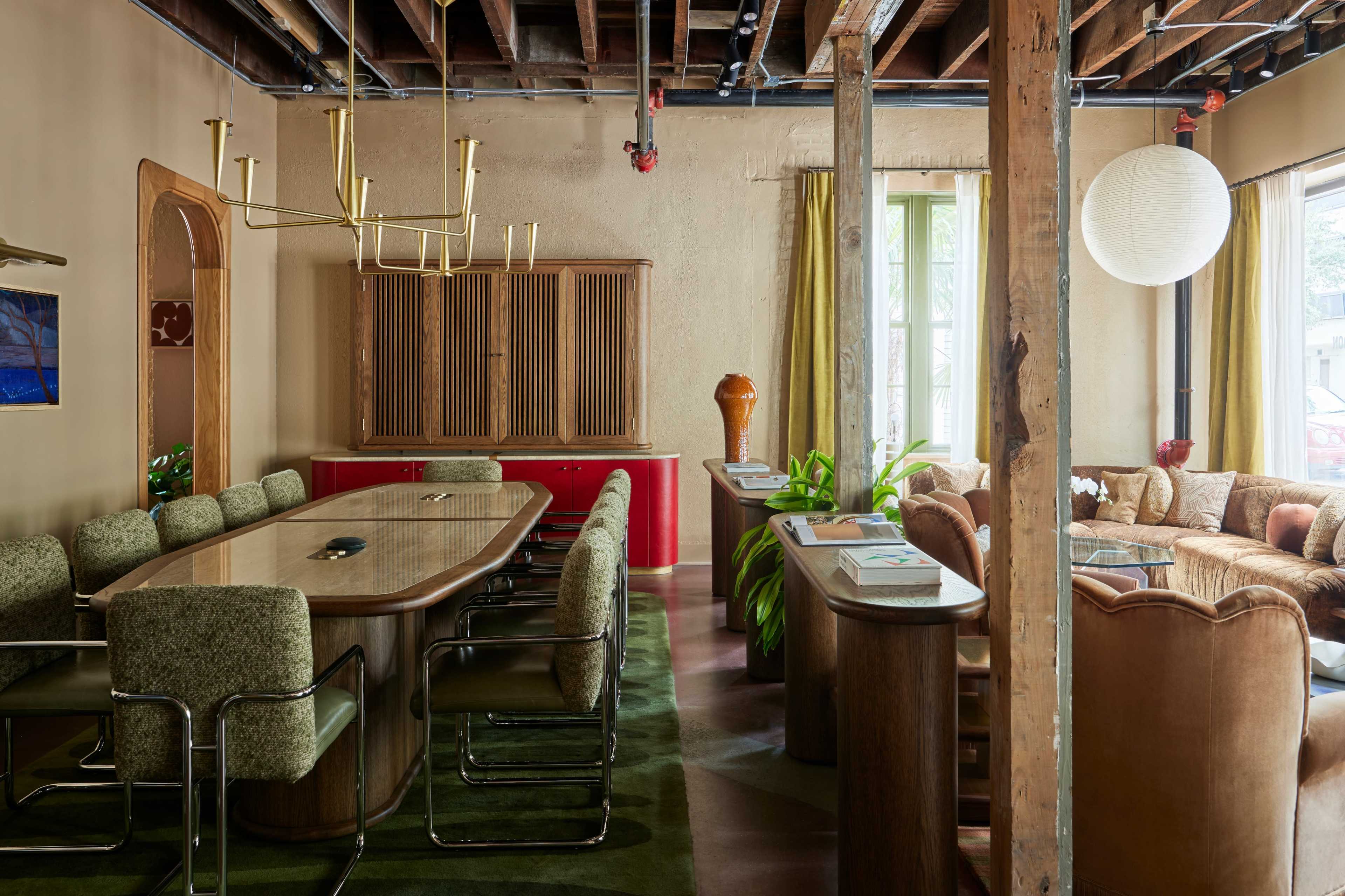 The image shows a modern interior space featuring a dining area with a large table surrounded by upholstered chairs, a wooden cabinet, and a cozy living area with a sofa, all under exposed ceiling beams.