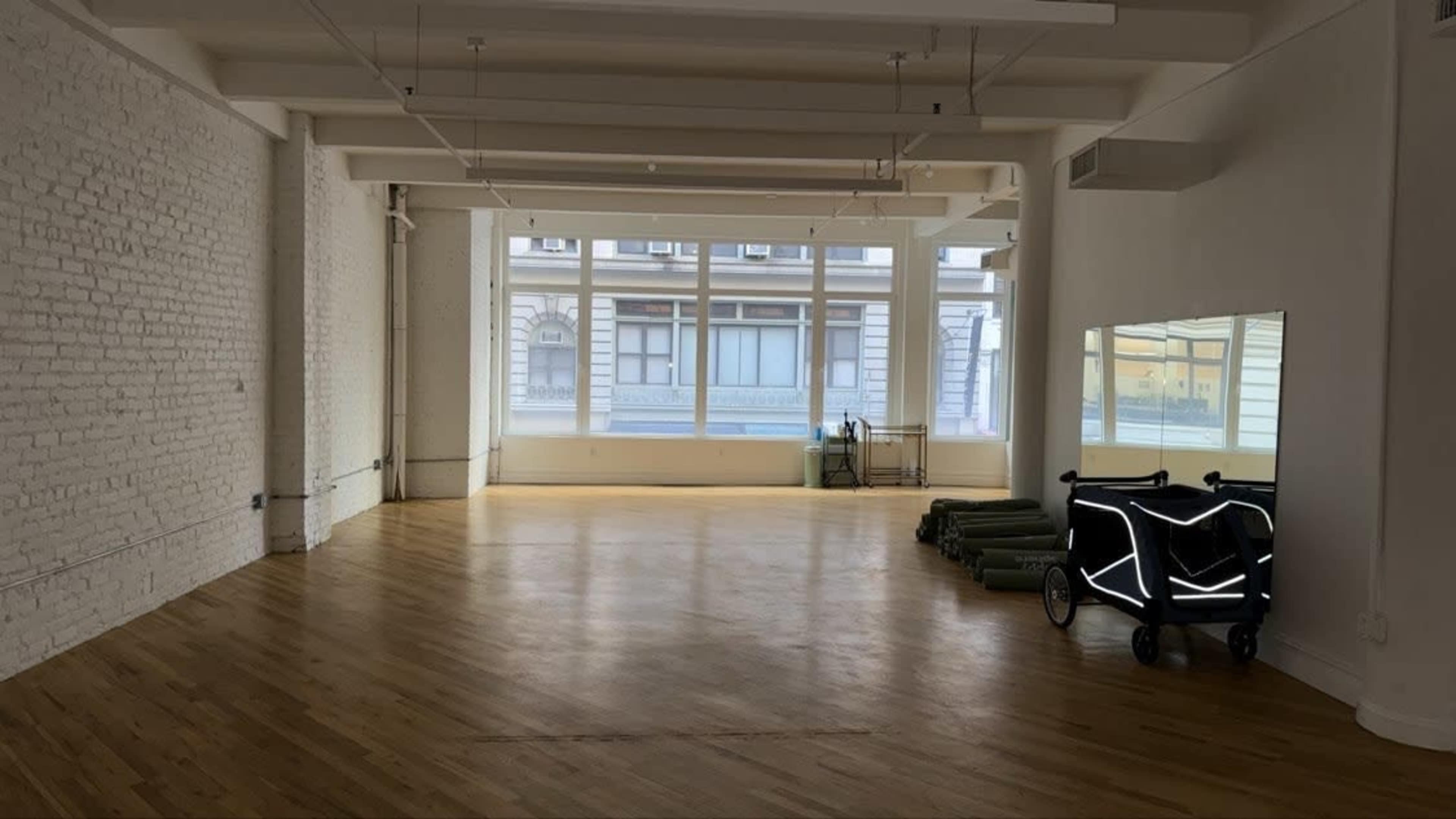 The image shows a spacious, empty room with wooden flooring and large windows, featuring a stroller with reflective details and rolls of green fabric stacked against the wall.