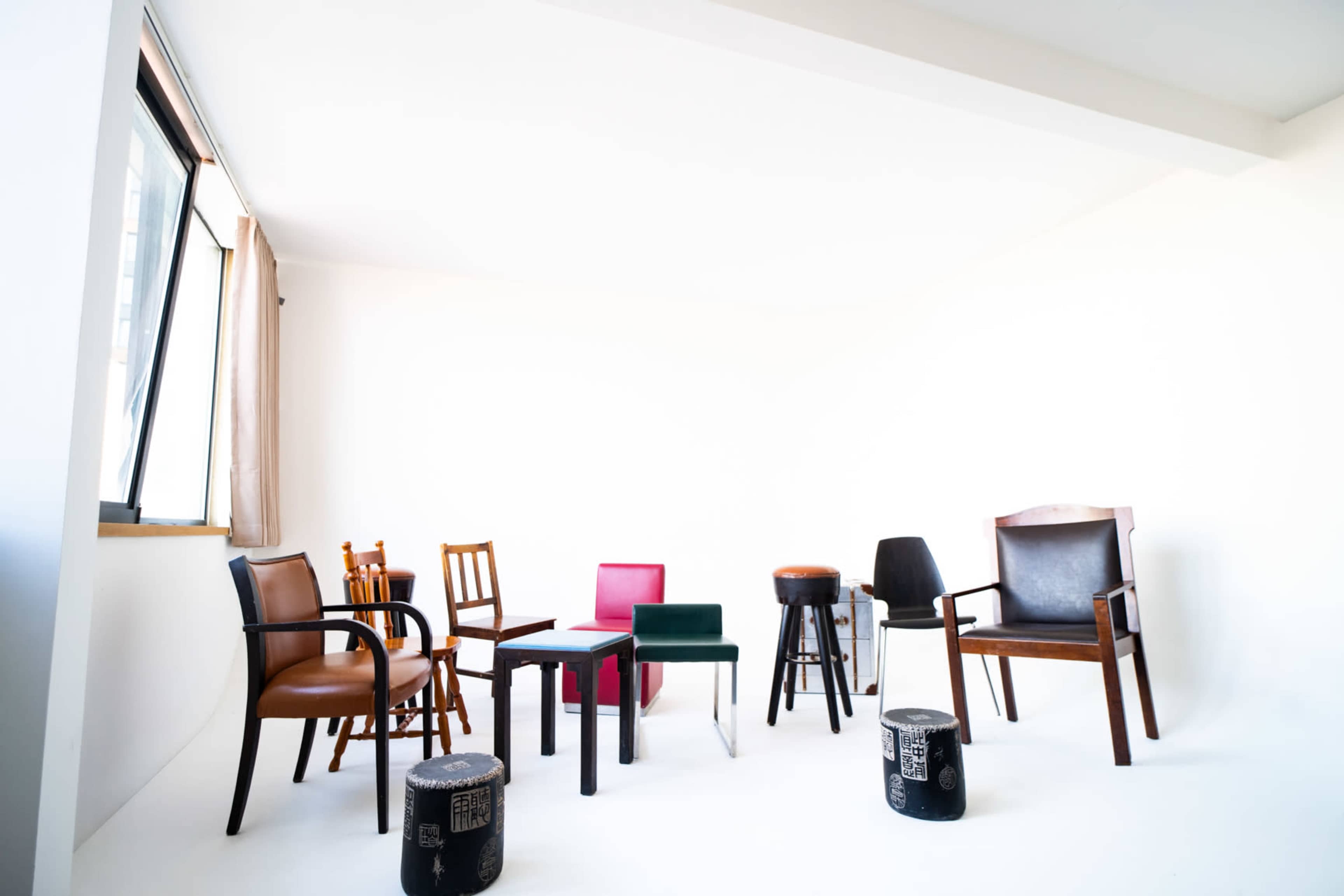 The image shows a variety of chairs and stools arranged in a white room with natural light coming through a window.