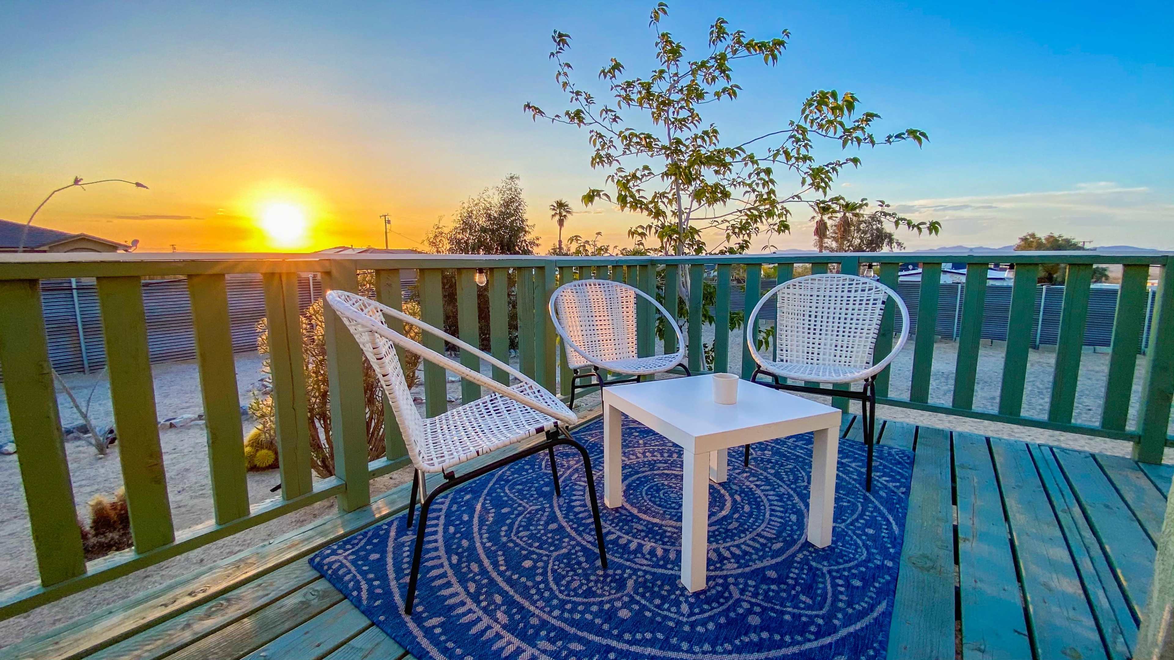 A small outdoor seating area features three wicker chairs surrounding a white table, with a sunset view in the background.