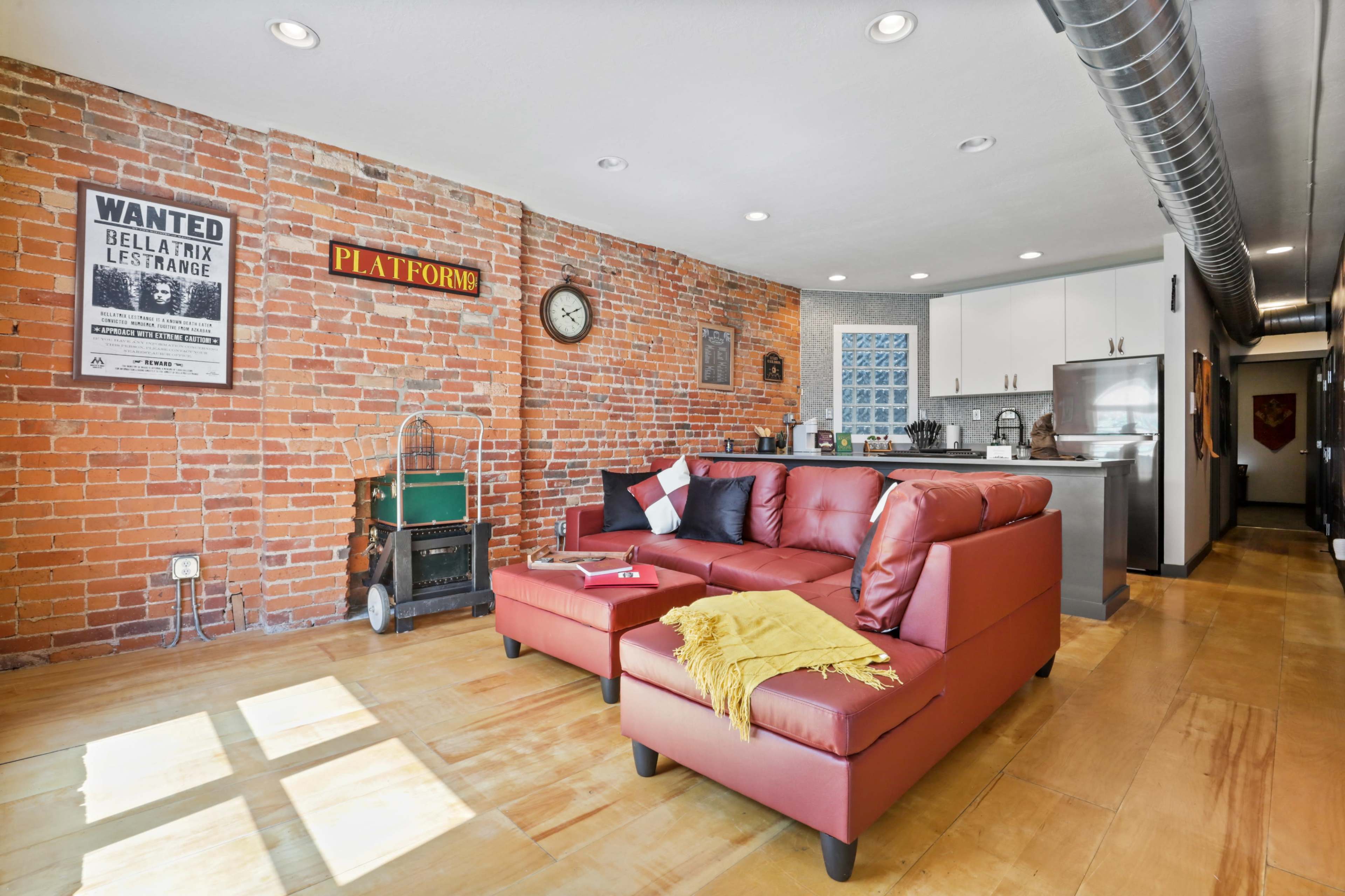 A modern living space with exposed brick walls, a red sectional sofa, a kitchen area, and various wall decorations.