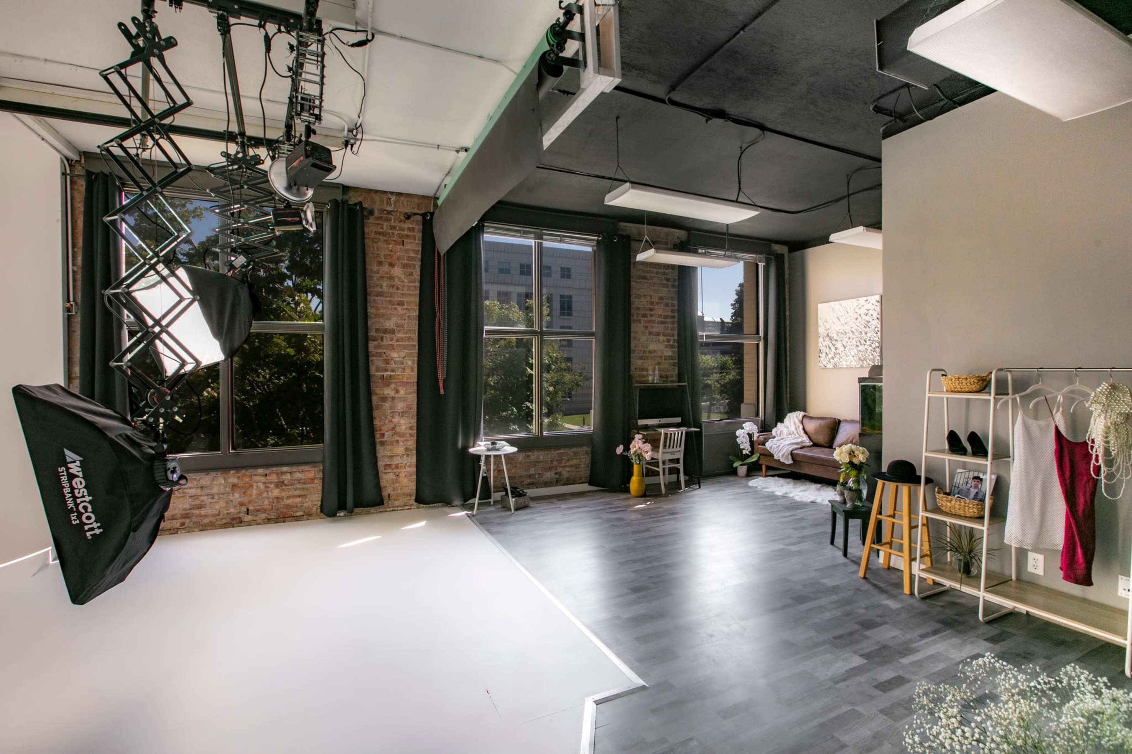 A well-lit studio space featuring a photography setup with softbox lights, a seating area, and a clothing rack against a backdrop of large windows.